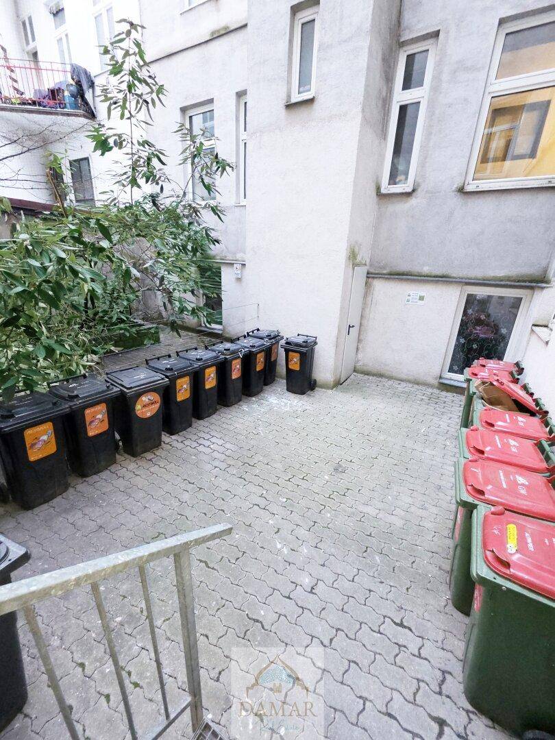 Außenbereich mit zahlreichen Mülltonnen und gepflastertem Boden, neben einer Gebäudefassade.