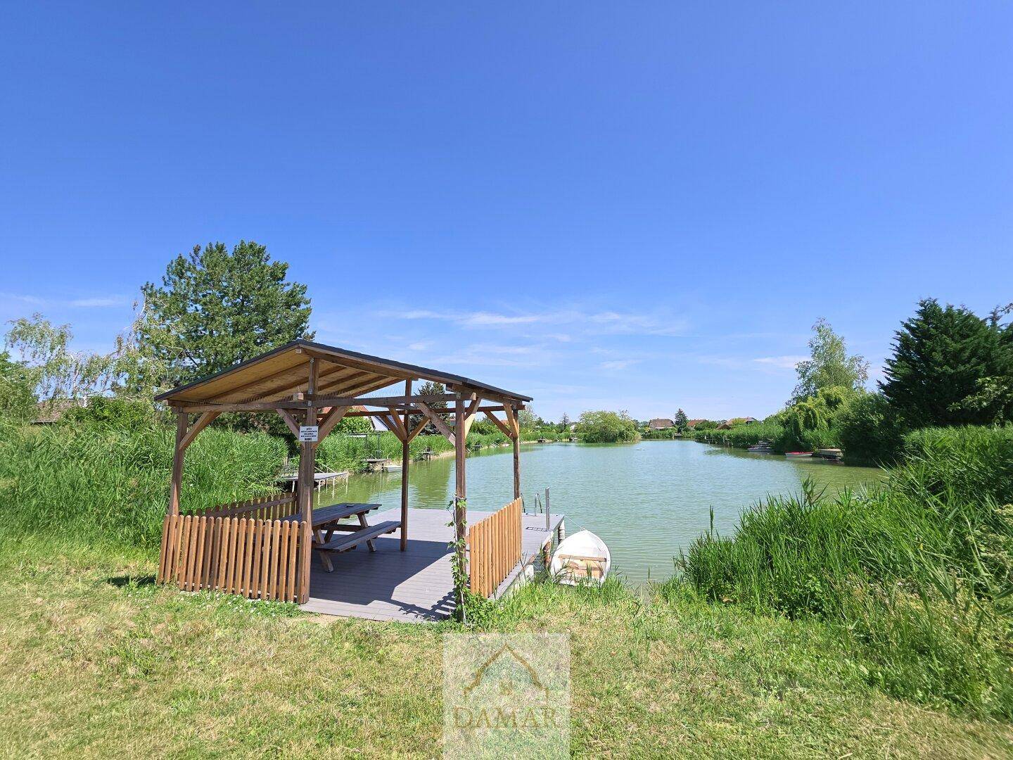 Holzpavillon mit Picknicktisch am Ufer eines ruhigen Sees unter blauem Himmel.
