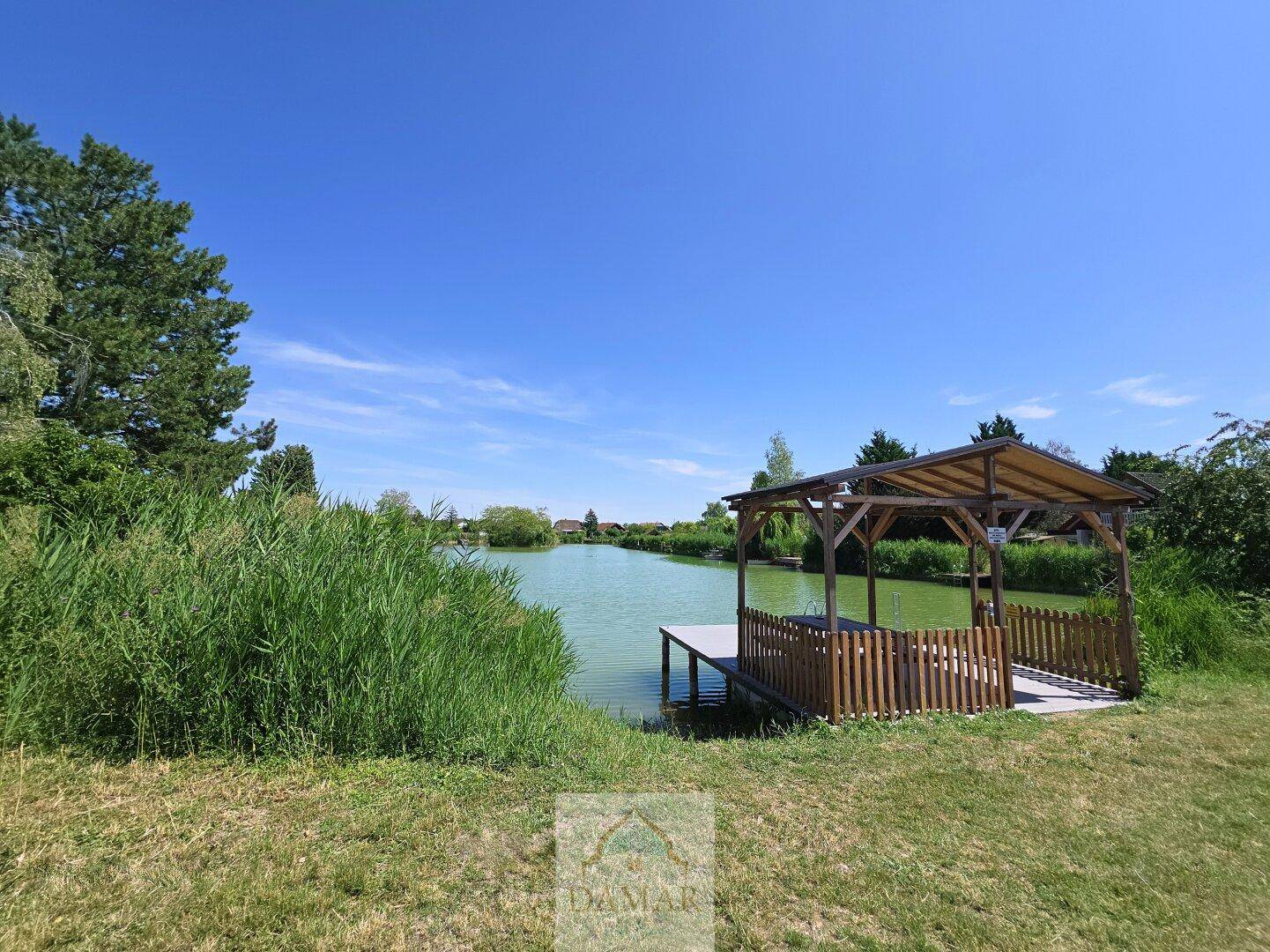 Holzpavillon am Seeufer, umgeben von Schilf und grüner Landschaft.