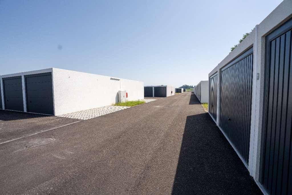 Eine Reihe moderner Garagenboxen mit dunklen Toren und asphaltierter Zufahrt unter einem klaren blauen Himmel.