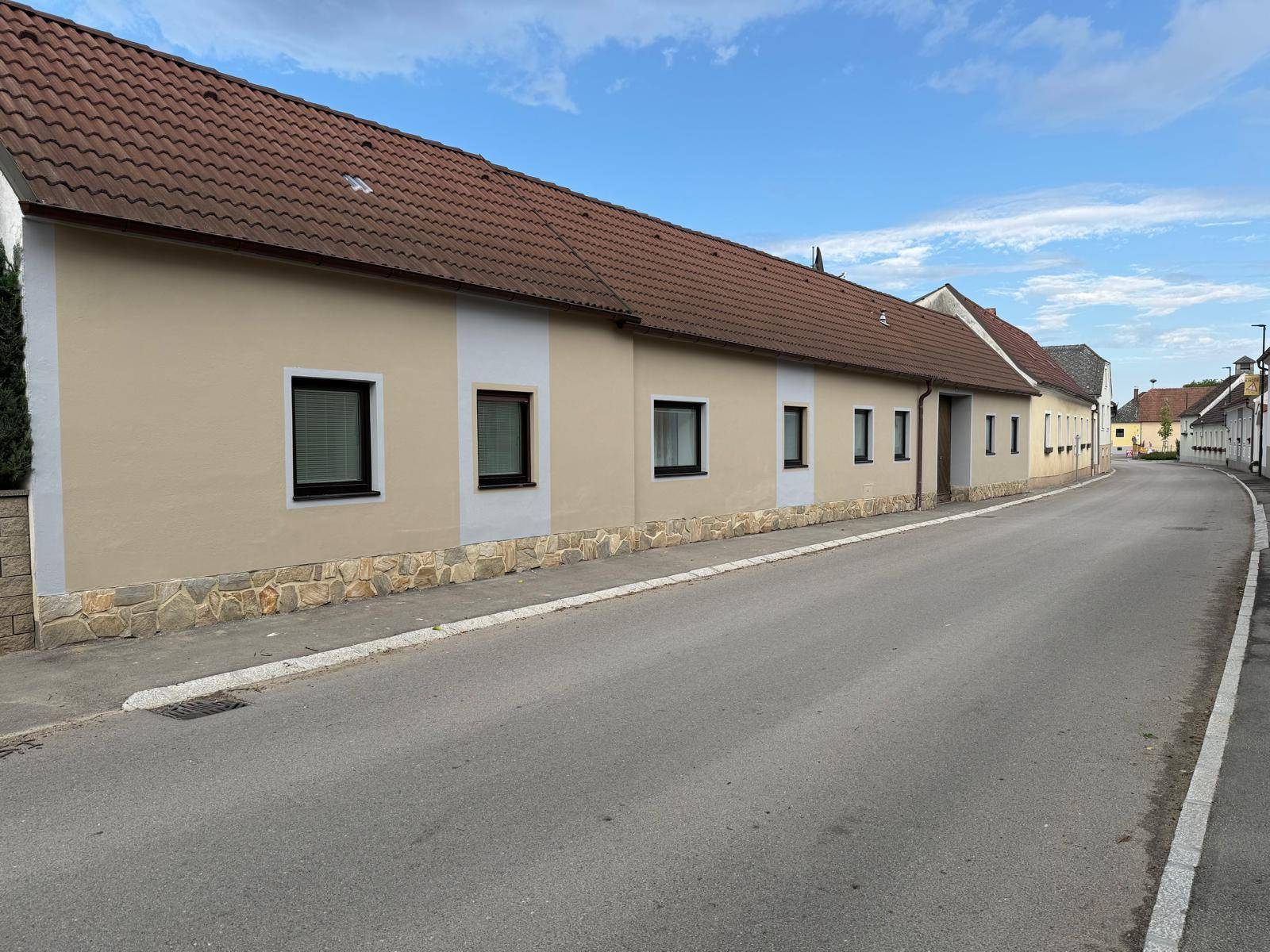 Lange Hausfassade mit mehreren Fenstern und beiger Putzfarbe entlang einer ländlichen Straße unter blauem Himmel.