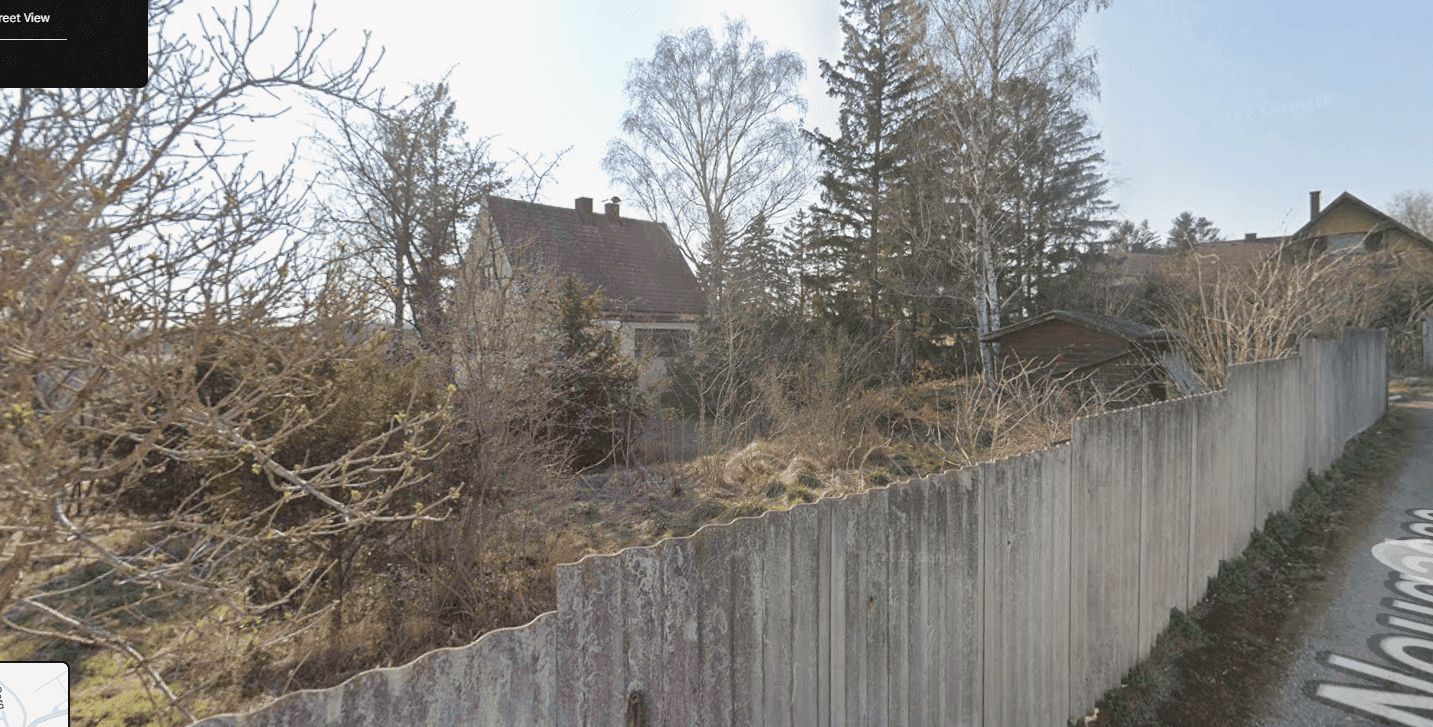 Blick auf ein verwildertes Grundstück mit Holzzaun und einem Haus mit rotem Dach im Hintergrund.
