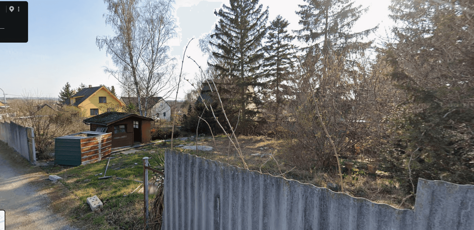 Weitläufiges Grundstück mit einem kleinen Gartenhaus, umgeben von Bäumen und einem Wellblechzaun, unter blauem Himmel.