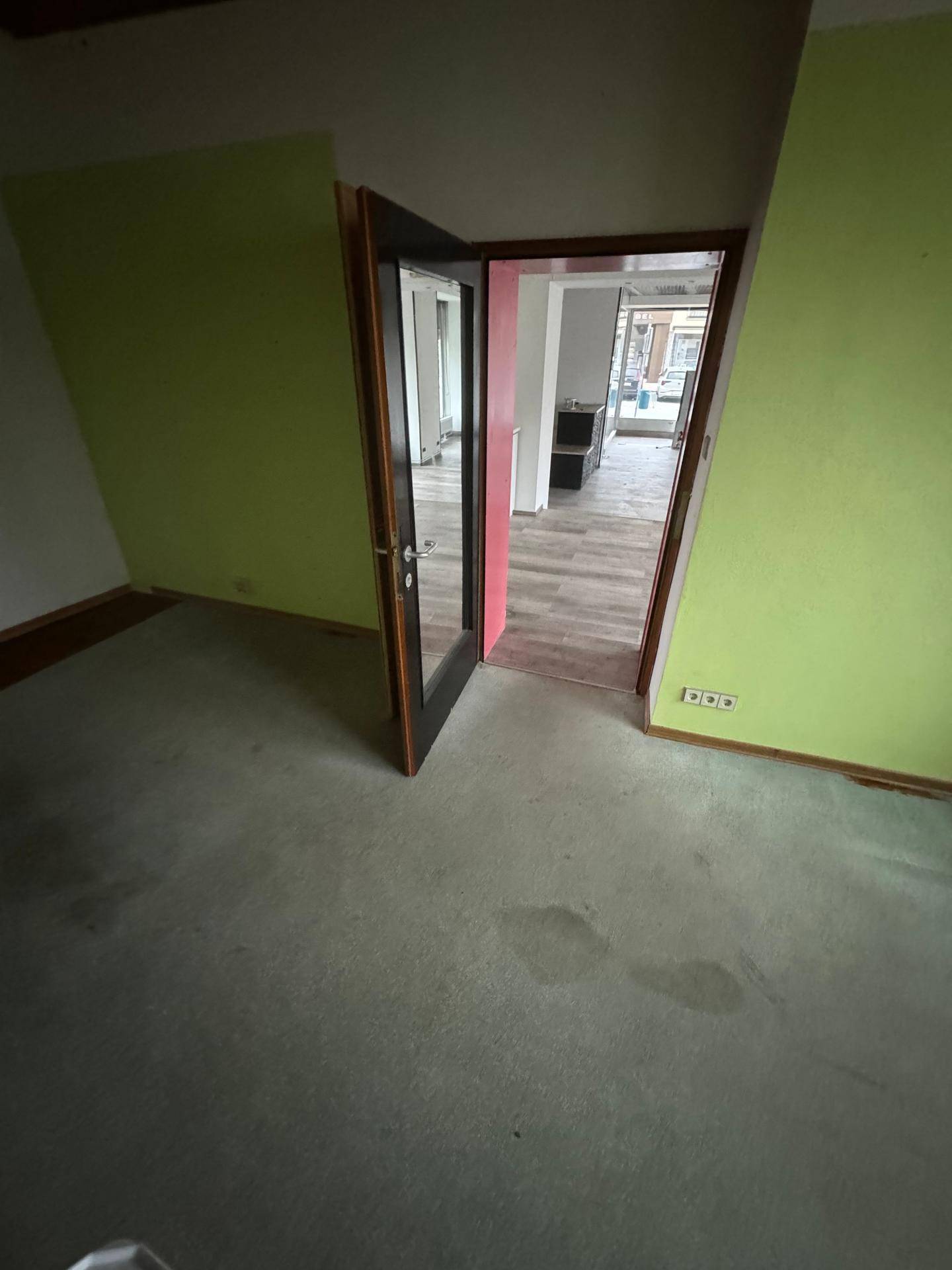 Raum mit grün gestrichenen Wänden und Teppichboden, mit Blick durch eine Glastür in den Ladenbereich.