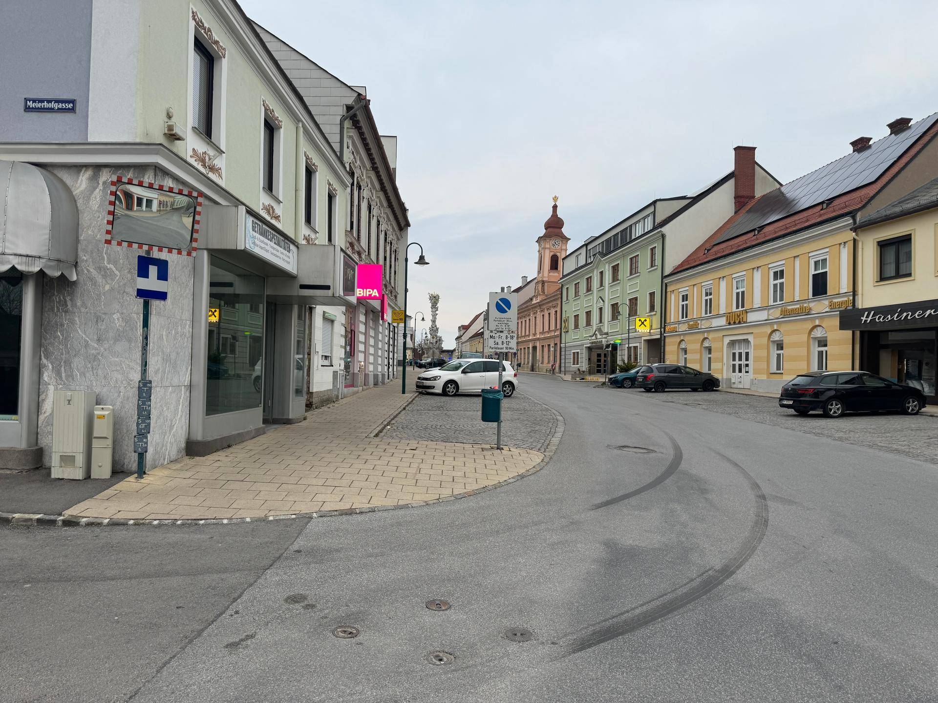 Eine belebte Straße in einer Stadt mit Geschäften, historischen Gebäuden und parkenden Autos.