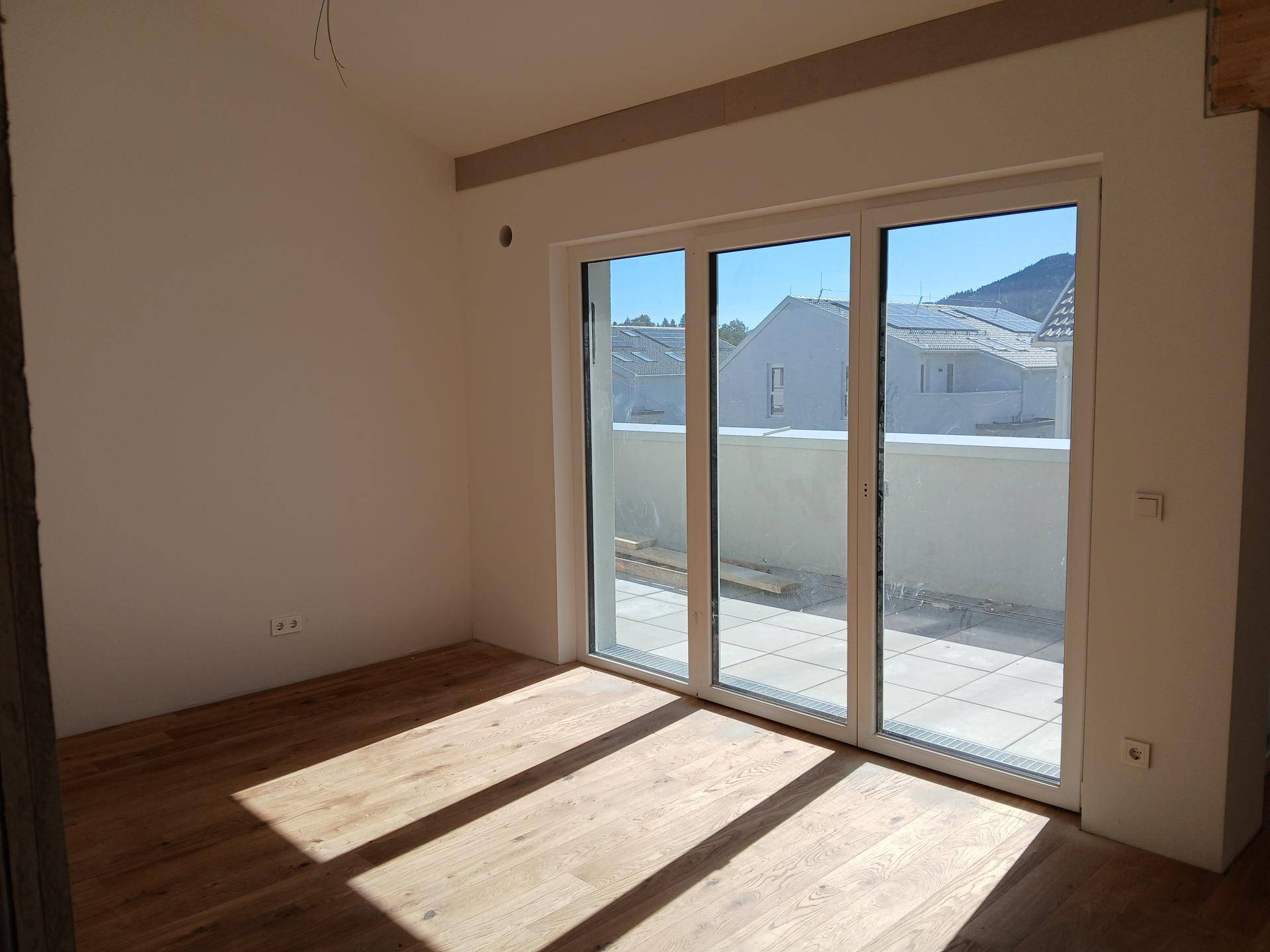 Unmöblierter Raum mit Holzboden und breiter Fensterfront, die viel Tageslicht hereinlässt.