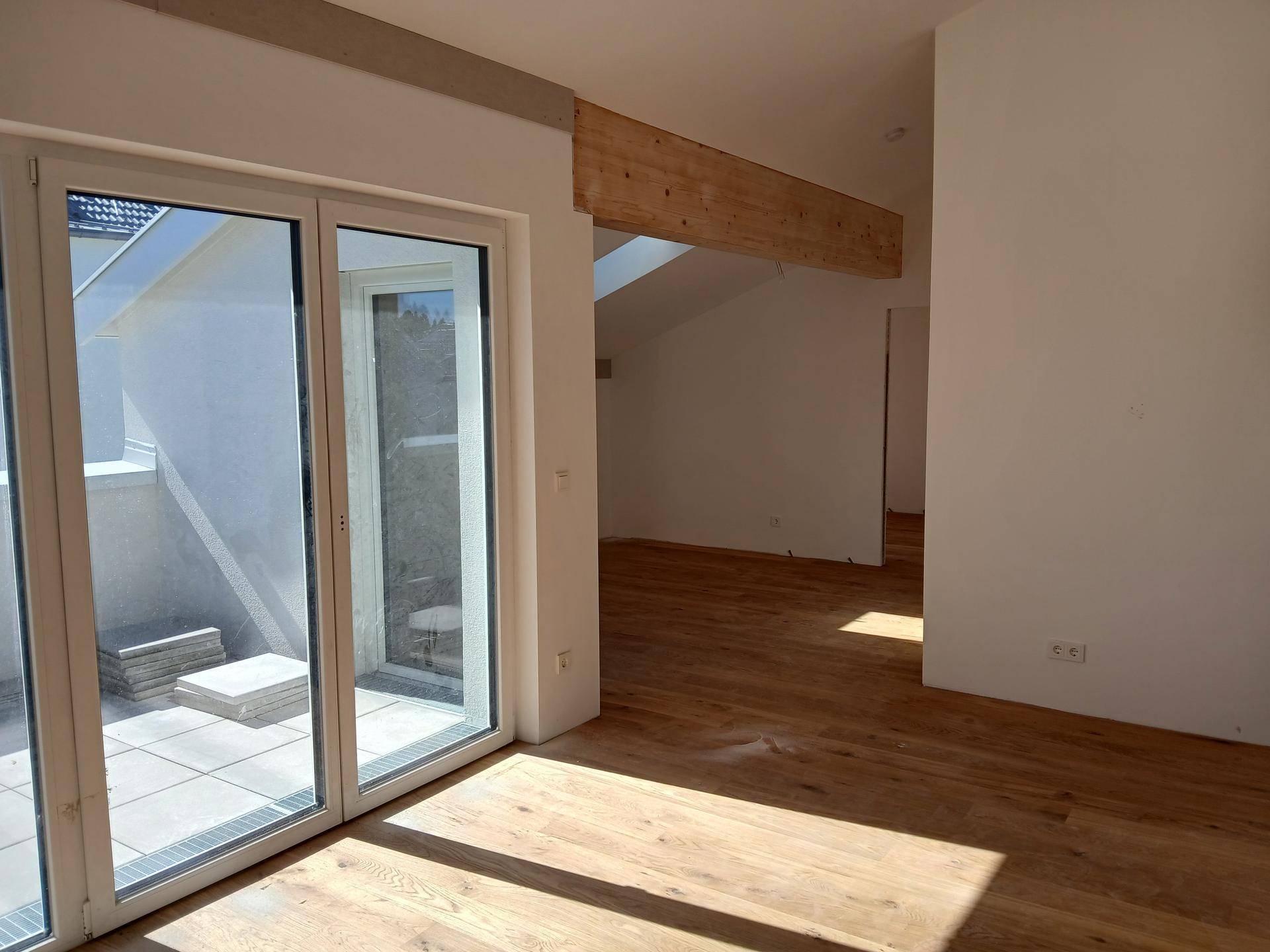 Lichtdurchfluteter Raum mit Holzboden und großen Fenstern, die auf eine Terrasse führen.