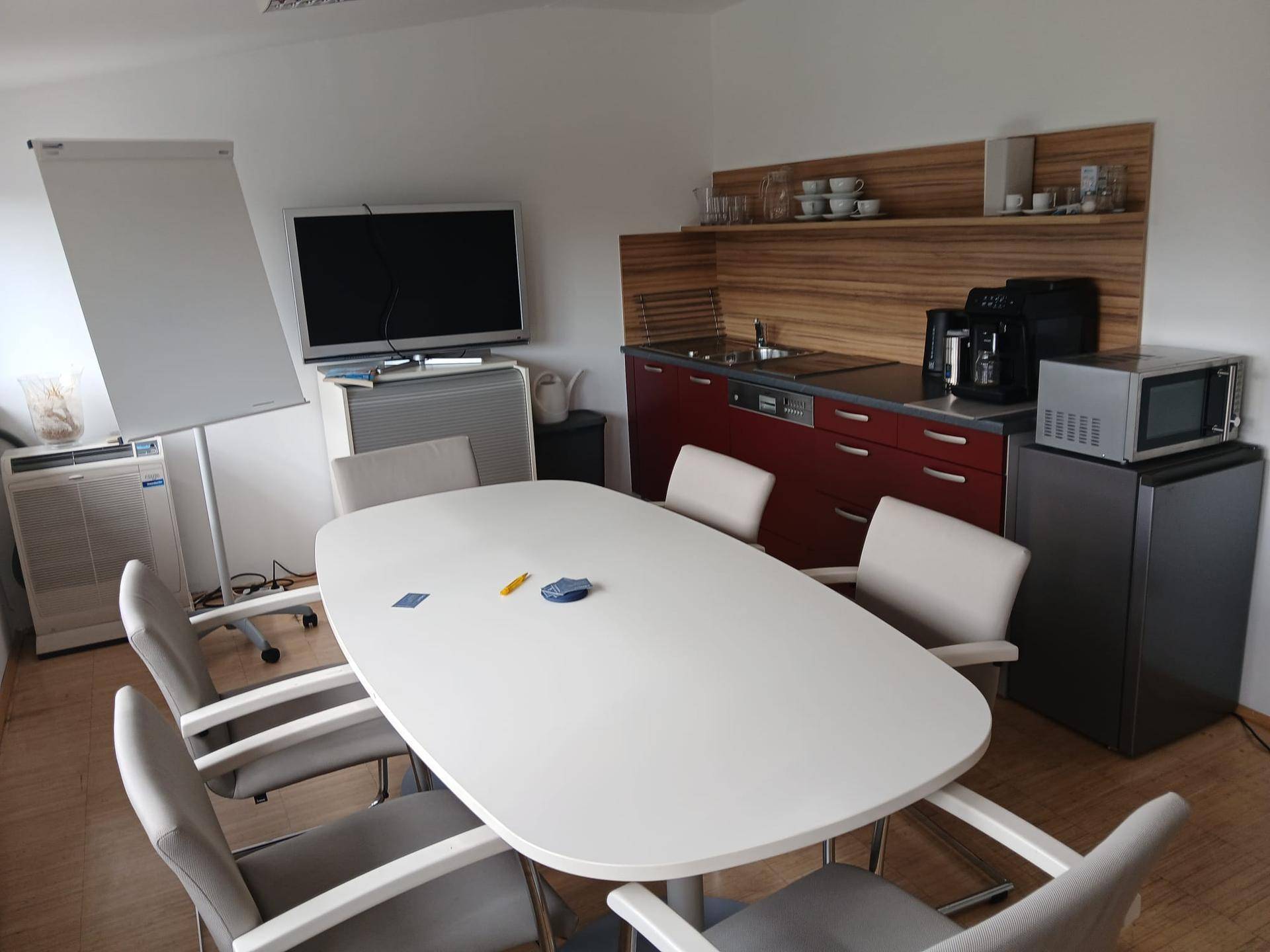 Konferenzraum mit großem Tisch, Stühlen, einem Fernseher und einer integrierten Küchenzeile.