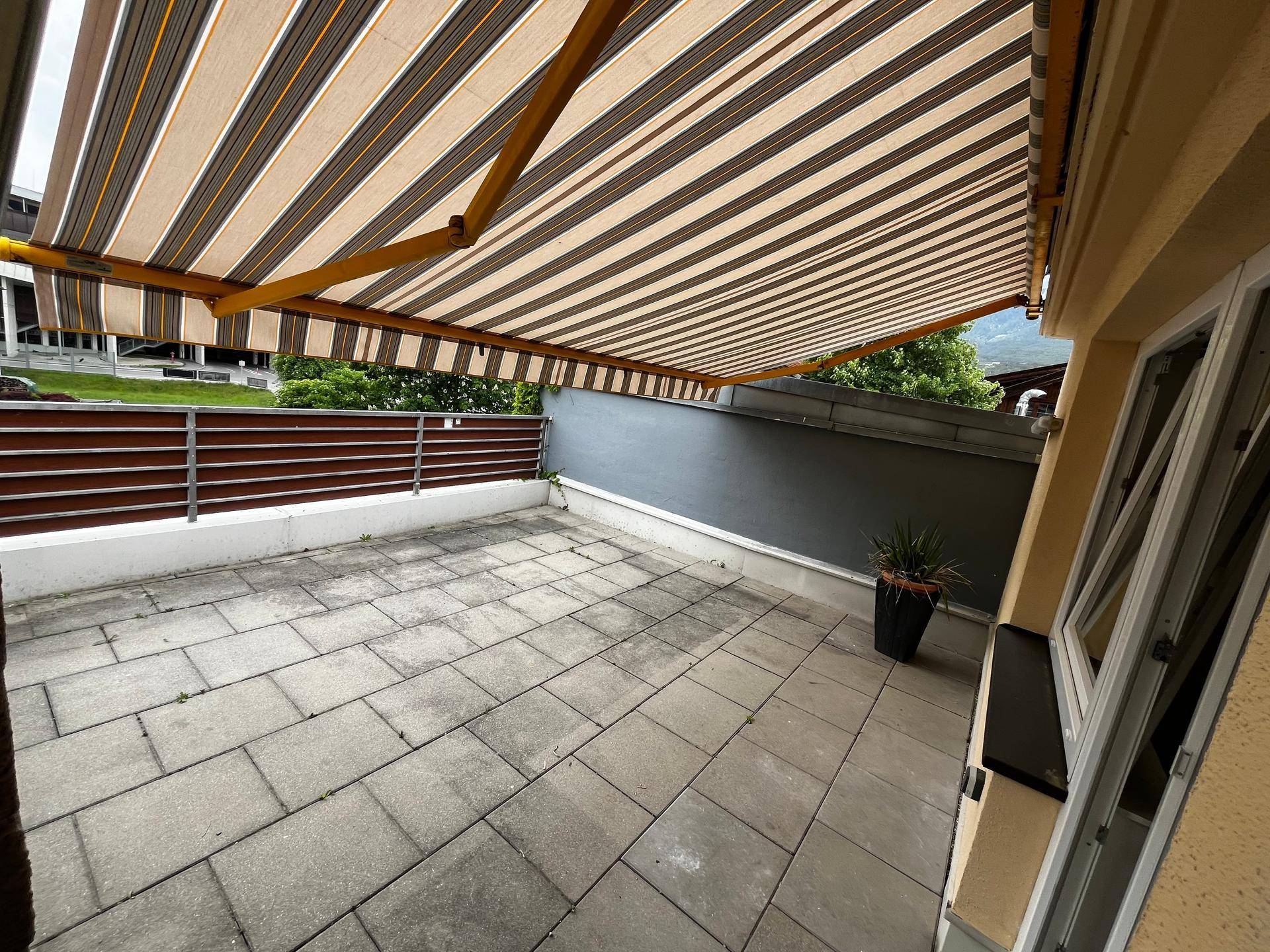 Geräumige Terrasse mit grauen Pflastersteinen, gestreifter Markise und Blick ins Grüne.