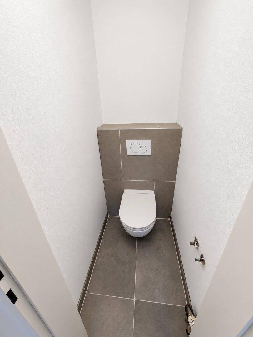 Separates WC mit Wand-WC und modernen grauen Fliesen, noch ohne Waschbecken.