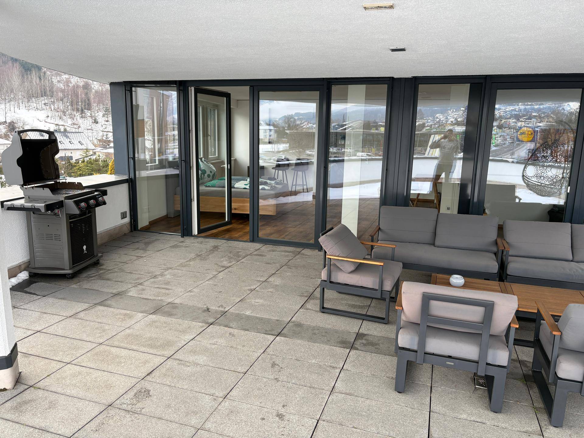 Helle Terrasse mit großzügigen Glasfronten zum Innenbereich, ausgestattet mit Lounge-Möbeln und Grill.