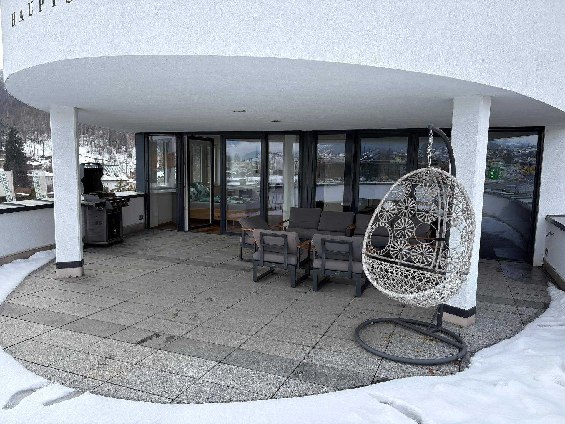 Geräumige überdachte Terrasse mit modernen Outdoor-Möbeln und einem Grill, umgeben von winterlicher Landschaft.