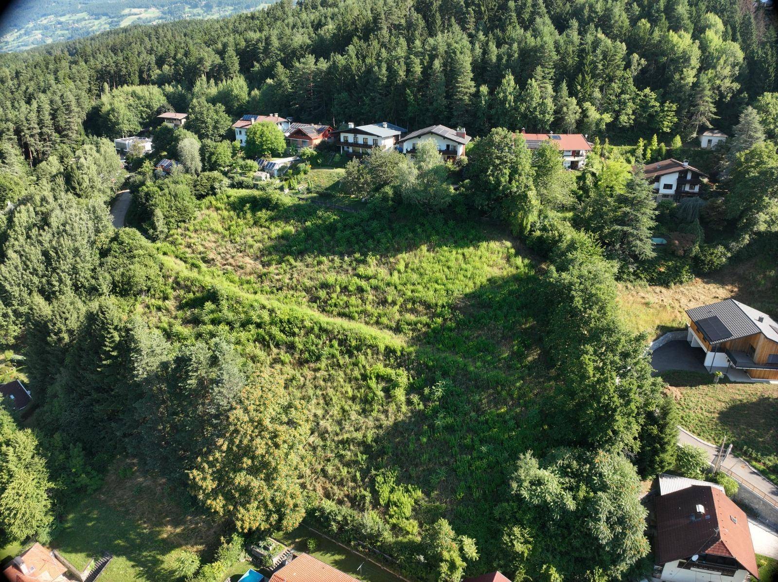 Luftaufnahme eines großen, grünen Baugrundstücks umgeben von Wohnhäusern und Waldgebieten.