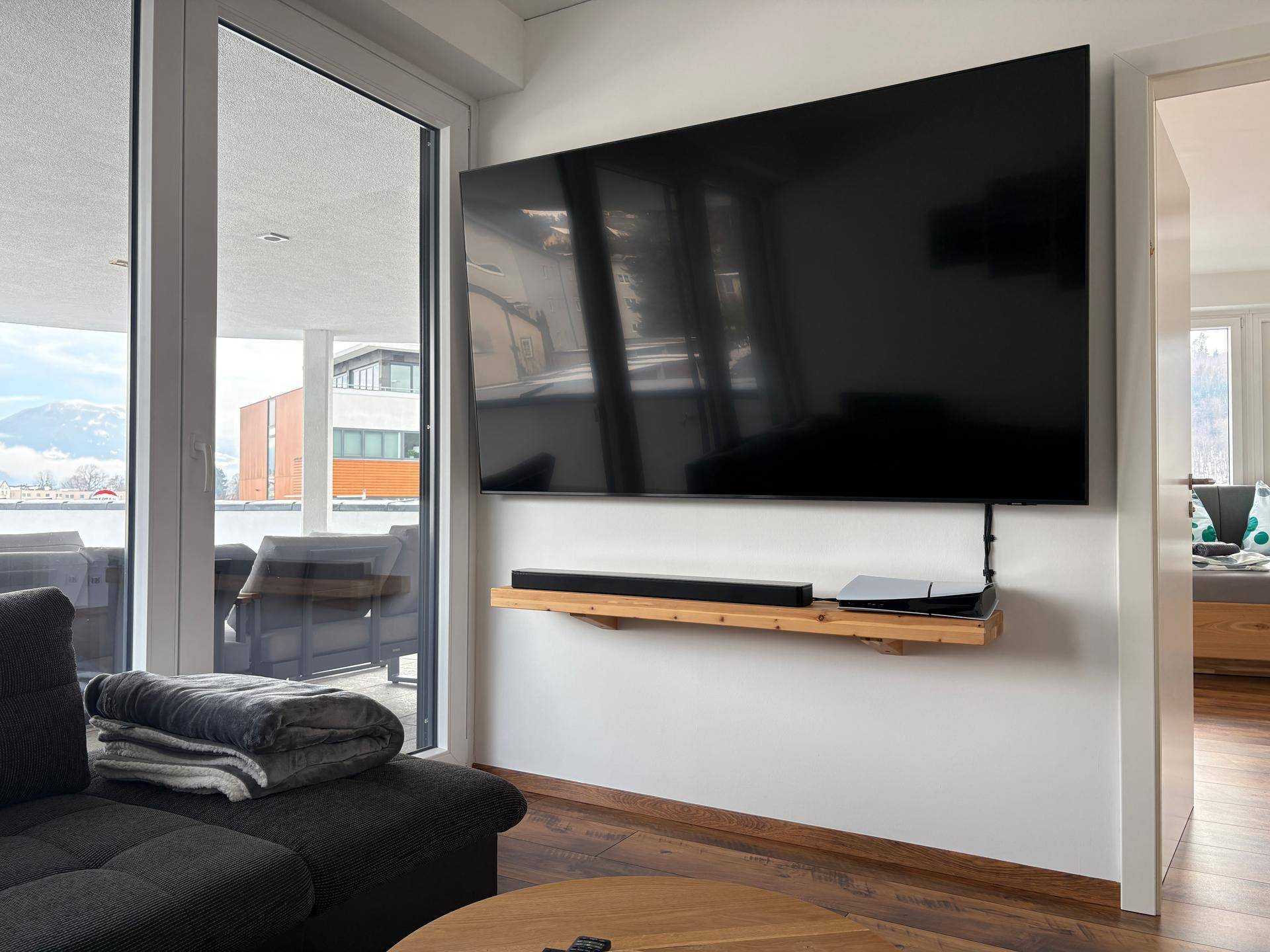 Wohnzimmer mit großem Wandfernseher, Soundbar und Zugang zur Terrasse durch expansive Glasfronten.