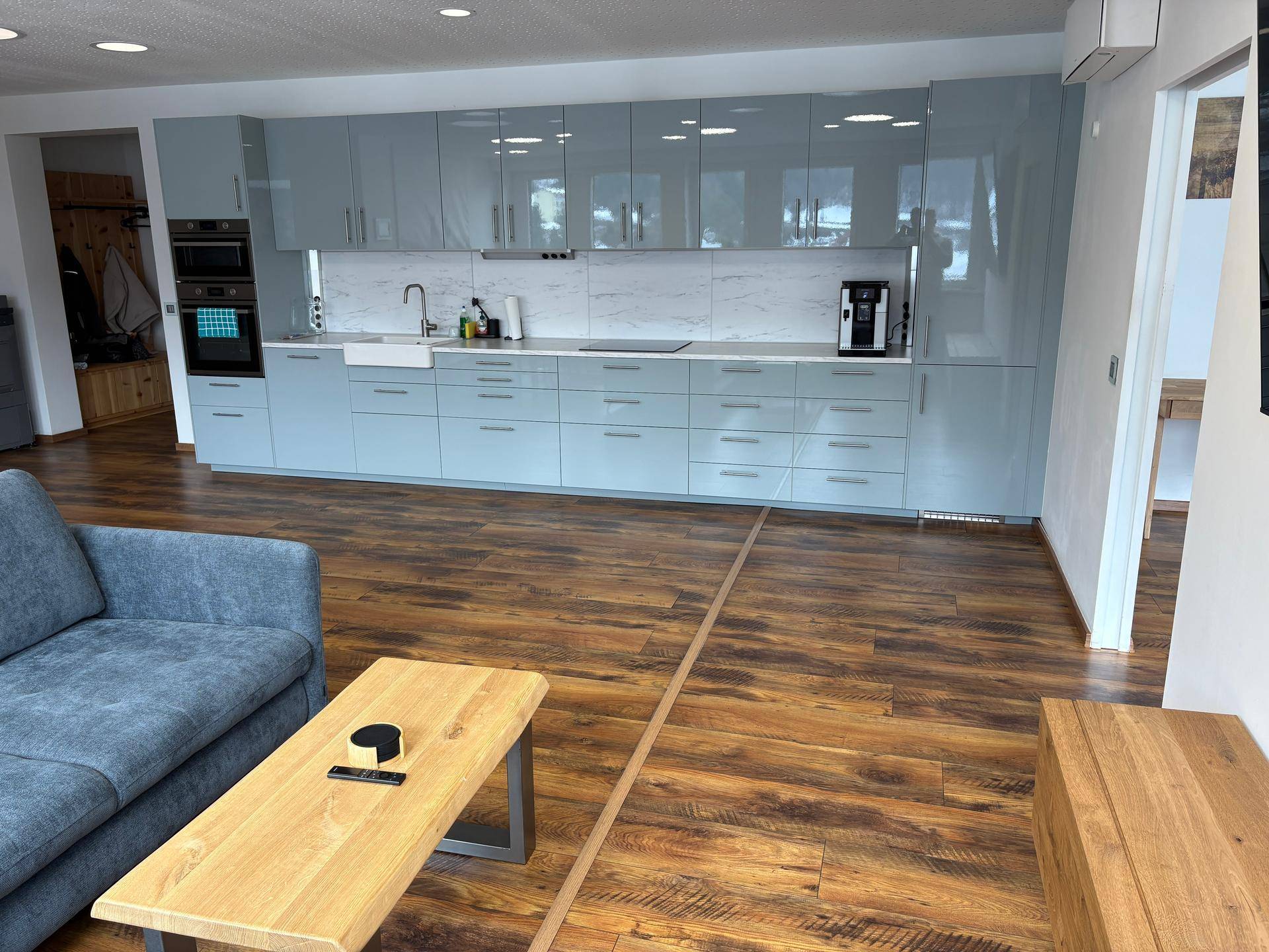 Moderne offene Küche mit hellblauen Hochglanzfronten, Marmor-Arbeitsplatte und Holzboden im Wohnbereich.