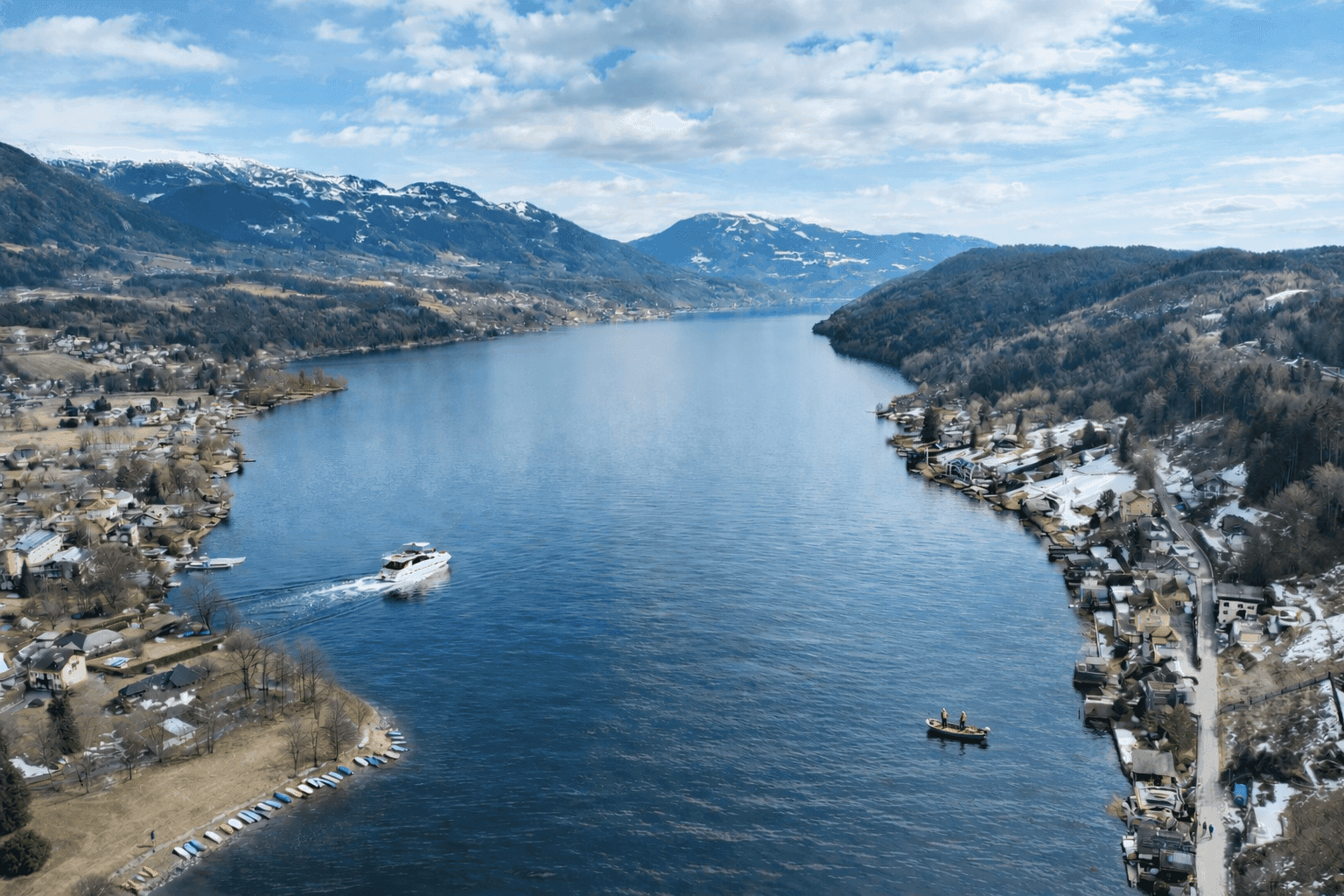 Panoramablick auf einen großen See, umgeben von schneebedeckten Bergen und Uferbebauung.