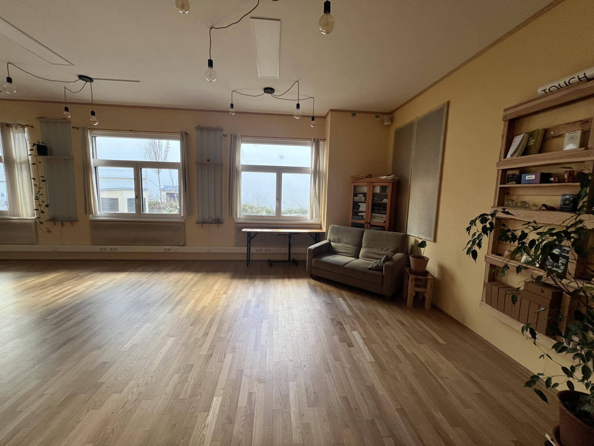 Heller Mehrzweckraum mit Holzfußboden, großen Fenstern und einer gemütlichen Couch, vielseitig nutzbar.