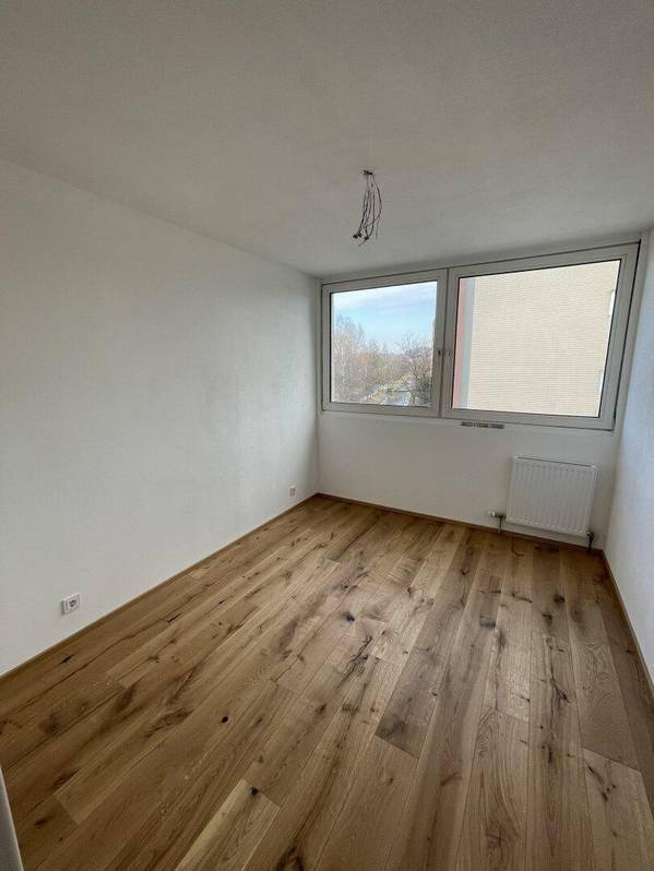 Helles Schlafzimmer mit Holzboden, weißen Wänden und einem Fenster mit Heizkörper.