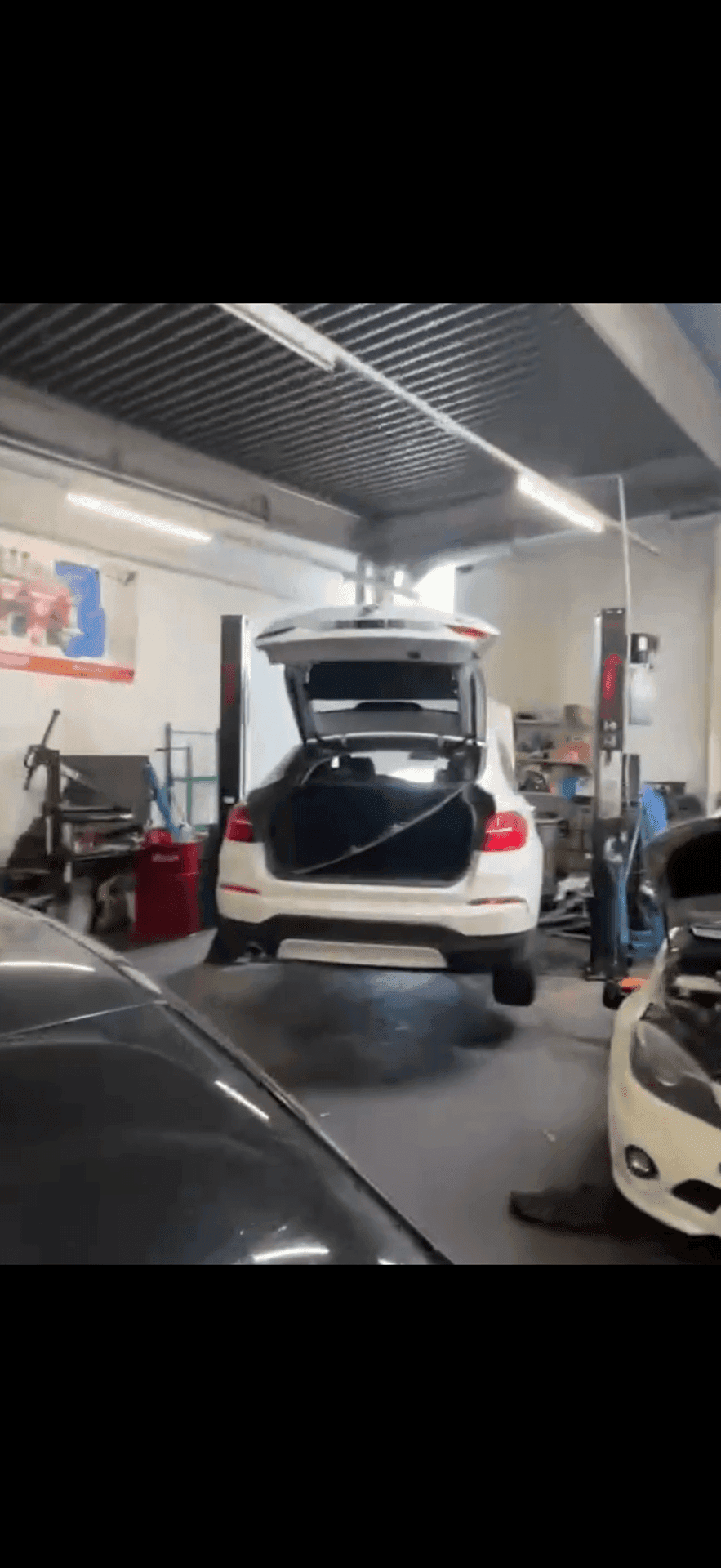 Blick in eine Autowerkstatt mit einem weißen Fahrzeug auf der Hebebühne und offenem Kofferraum.
