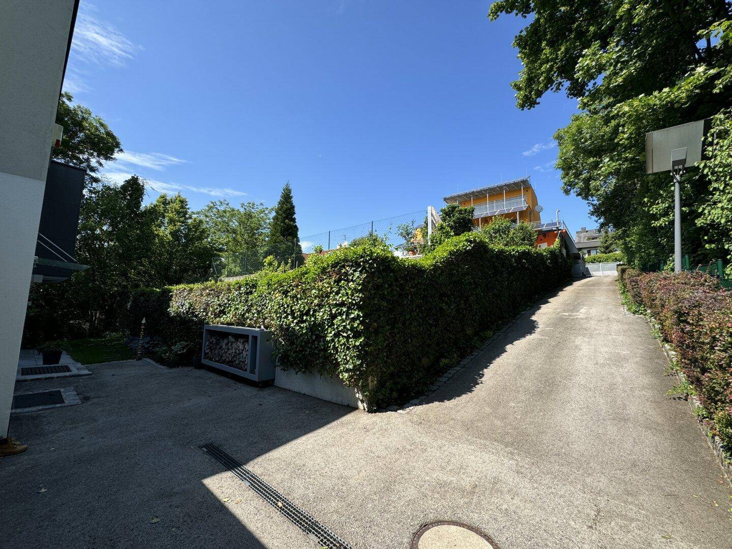 Zufahrt zum Grundstück mit Basketballkorb und grüner Hecke unter blauem Himmel.