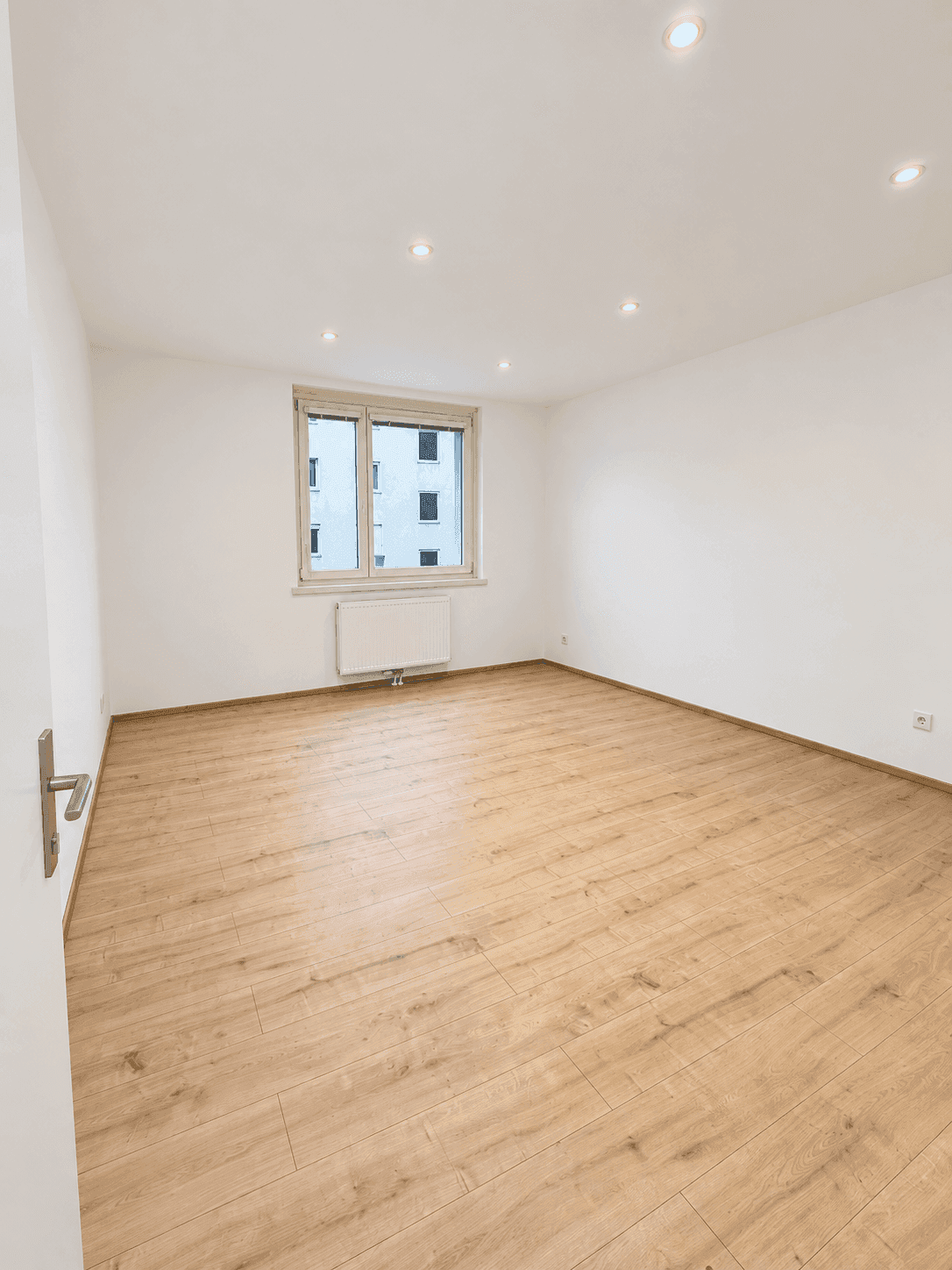 Helles, leeres Zimmer mit Laminatboden und einem Fenster mit Blick auf andere Gebäude, ideal für individuelle Gestaltung.