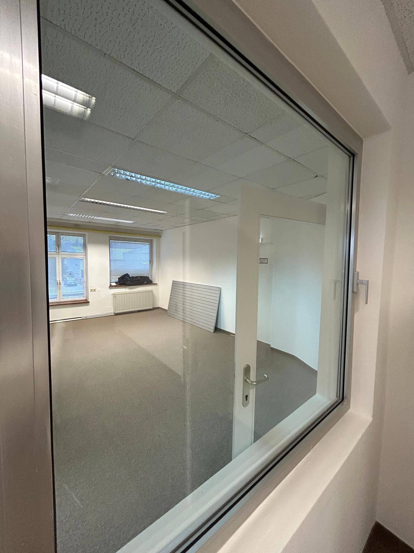 Blick durch eine Glaswand in ein leeres Büro mit Teppichboden und abgehängter Decke.