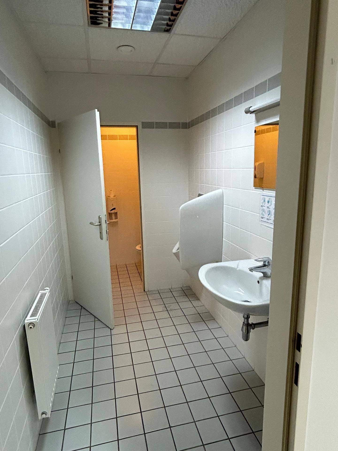 Gefliestes Badezimmer mit Urinal, Waschbecken und Zugang zu einer weiteren Toilette.