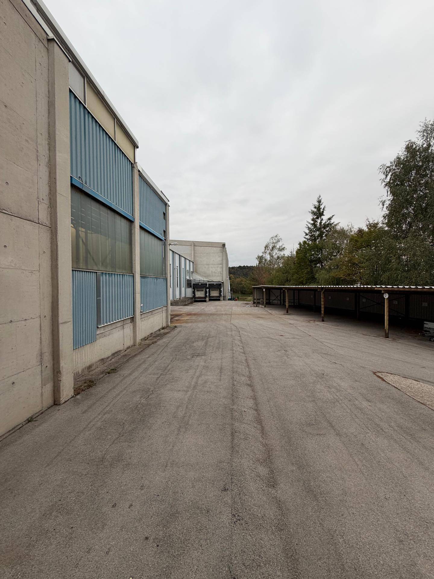 Außenansicht eines Industriegebäudes mit Betonfassade und blauer Verkleidung, daneben ein überdachter Parkplatz.