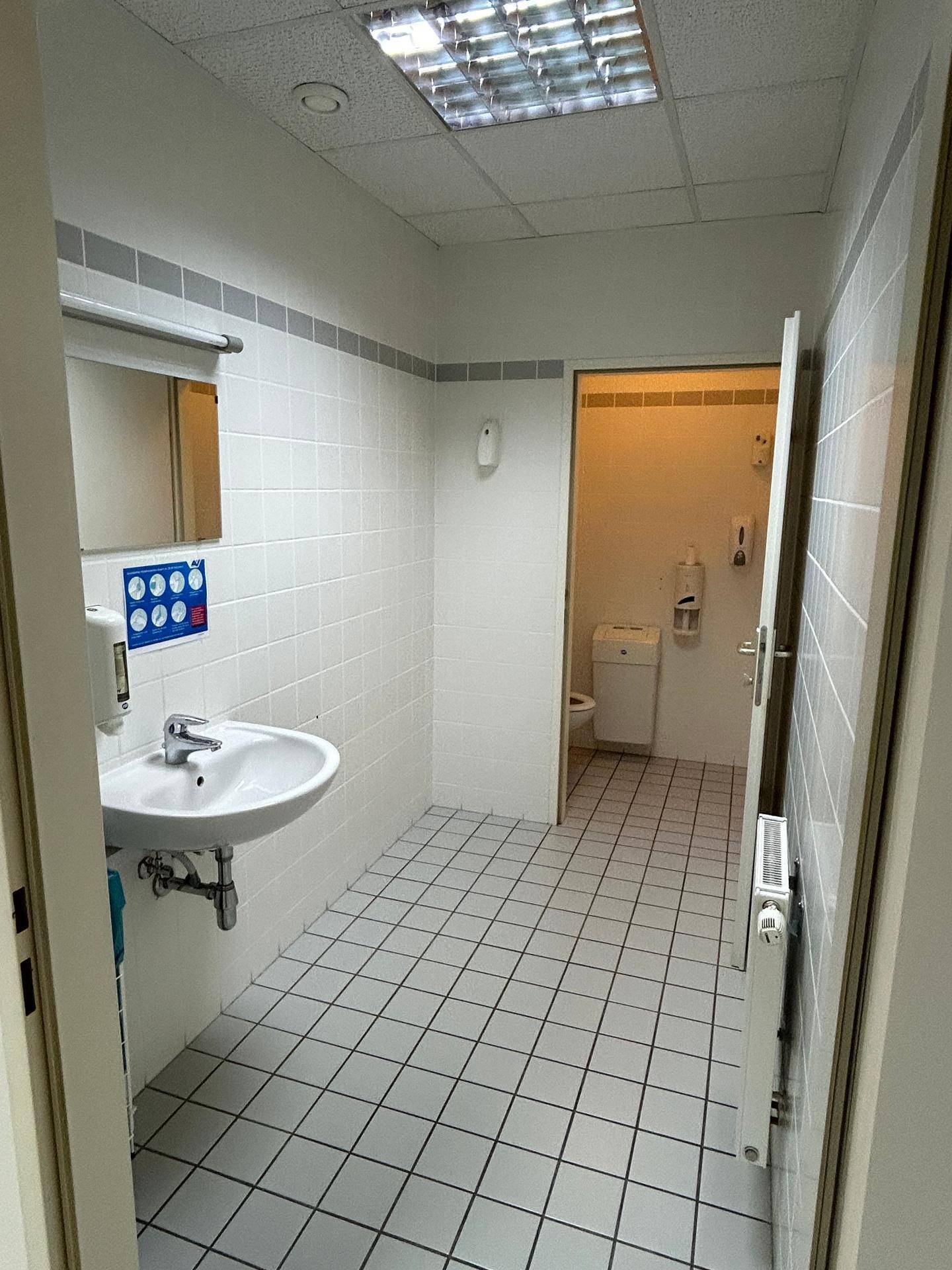 Gefliestes Badezimmer mit Waschbecken, Spiegel und Zugang zu einer weiteren Toilette.