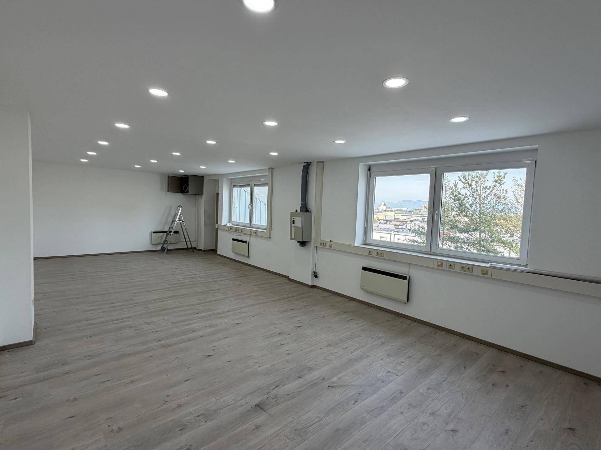 Geräumiger Büroraum mit mehreren Fenstern, die viel Tageslicht hereinlassen, und hellem Laminatboden.