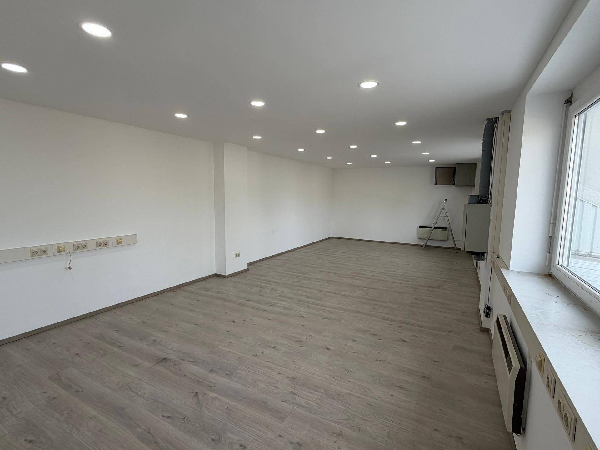 Heller, leerer Büroraum mit modernem Laminatboden, weißen Wänden und vielen Deckenstrahlern.