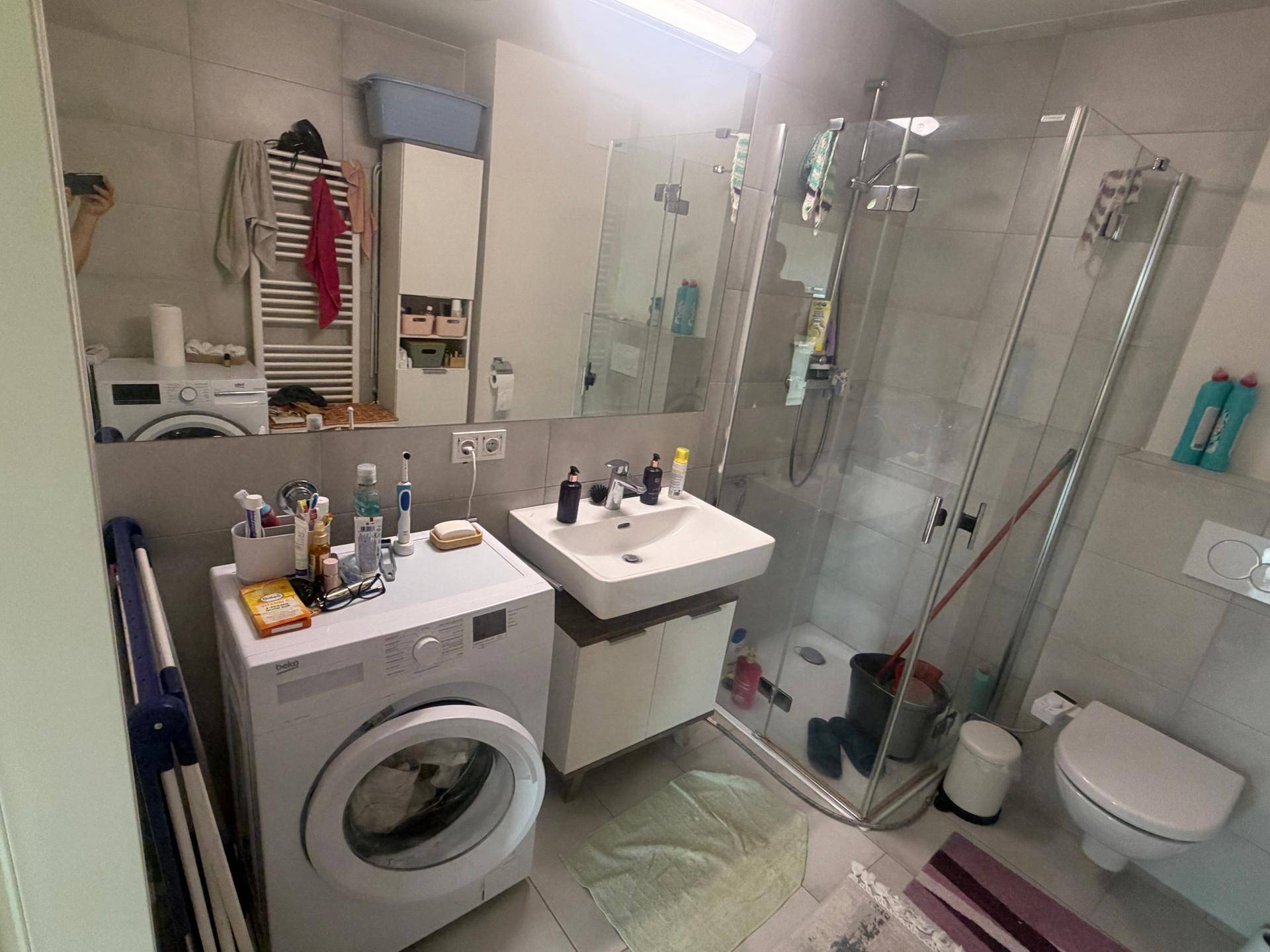 Modernes Badezimmer mit grauen Fliesen, Duschkabine, Waschbecken und Waschmaschine.