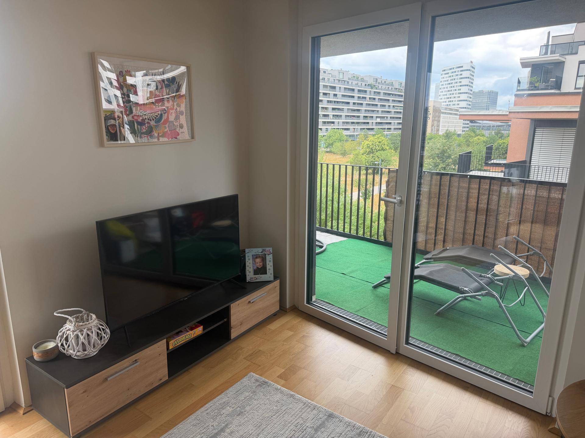 Heller Wohnbereich mit Fernseher und großem Fenster, das auf einen Balkon mit Stadtblick führt.