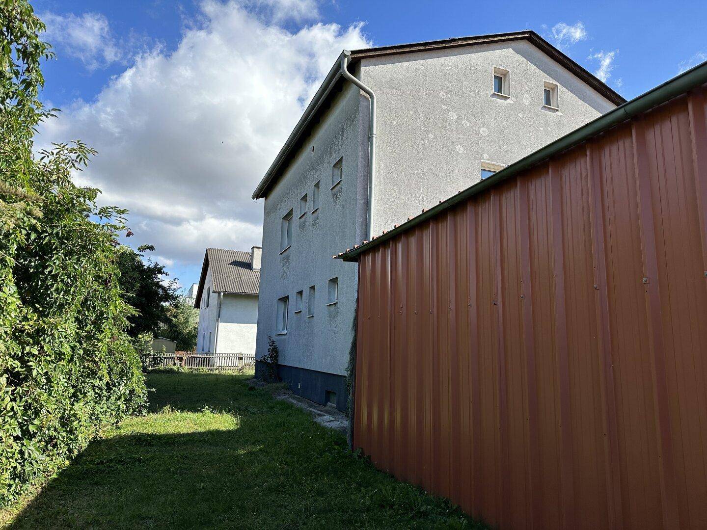 Seitenansicht des Hauses und eines Schuppens mit Rasenfläche und Hecke unter blauem Himmel.