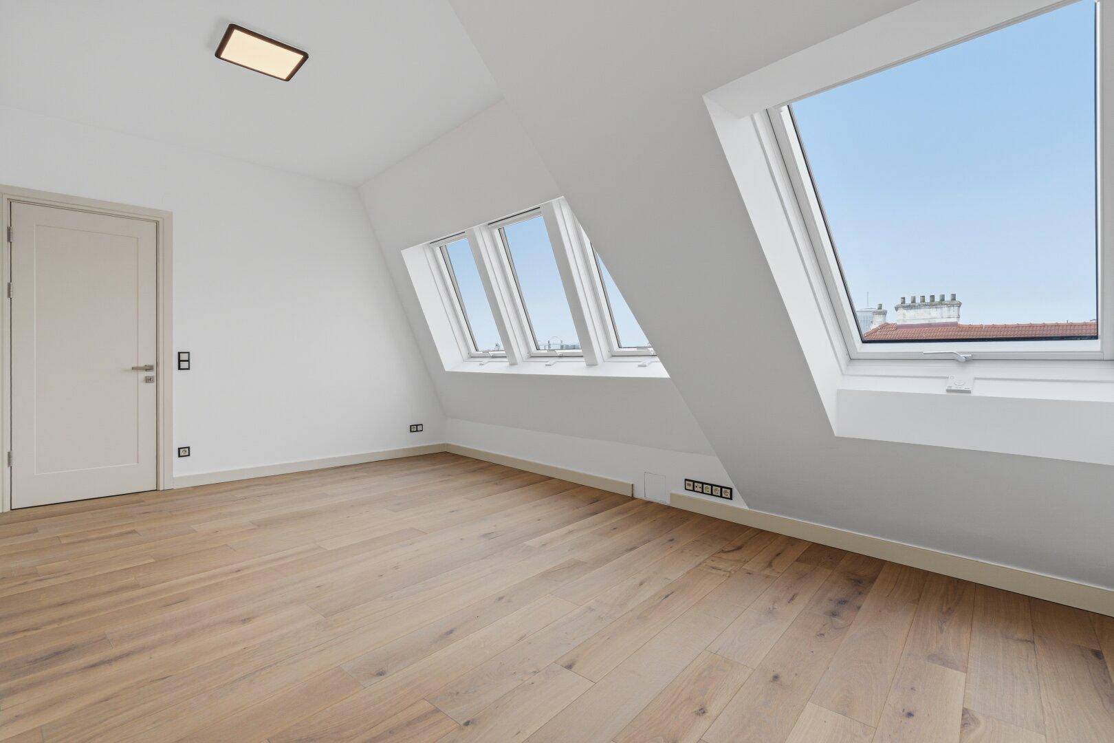 Helles, leeres Schlafzimmer mit Holzboden, Dachschrägenfenstern und weißer Tür.