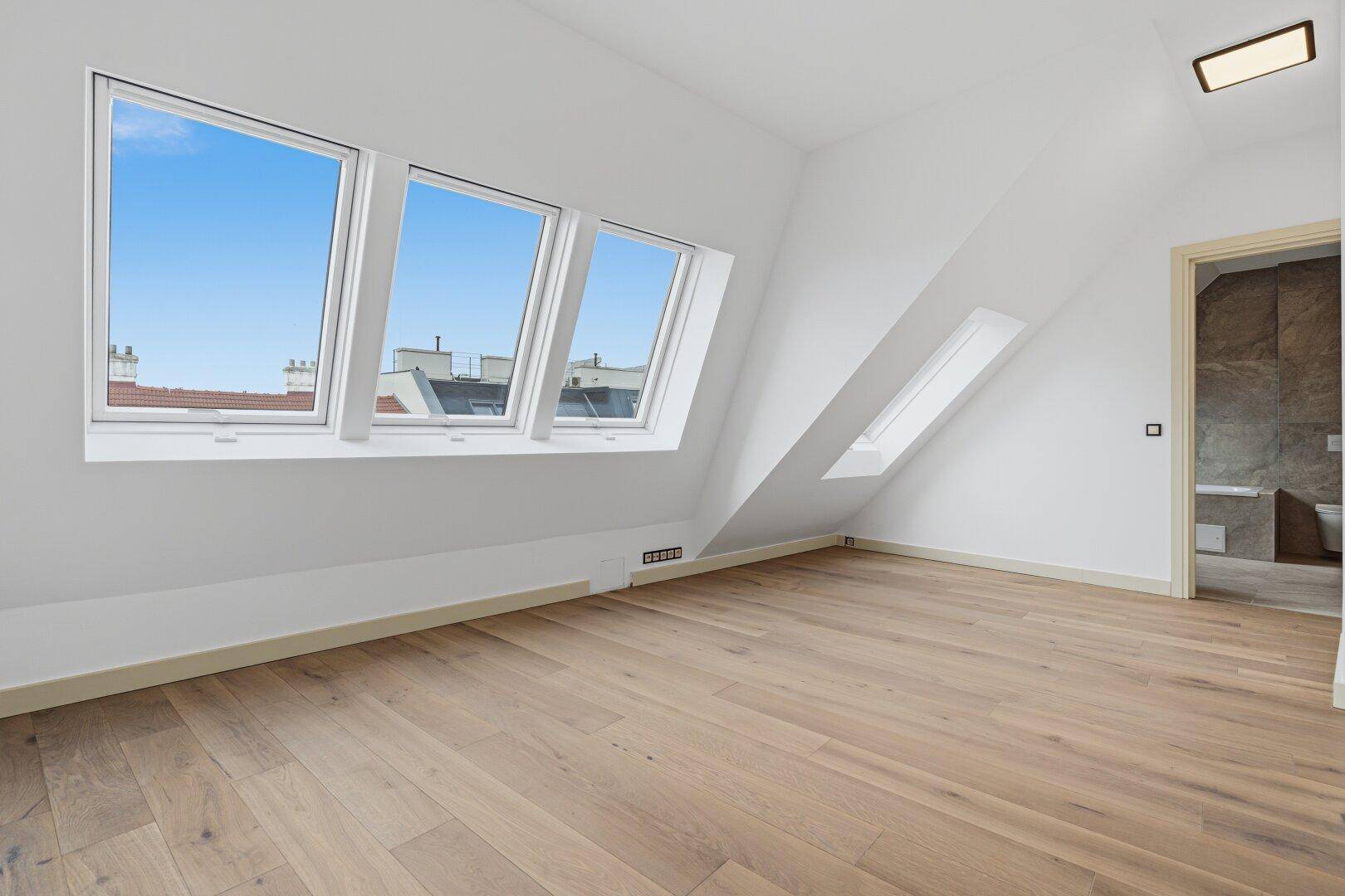 Helles, leeres Schlafzimmer mit Holzboden, Dachschrägenfenstern und Zugang zum Bad.