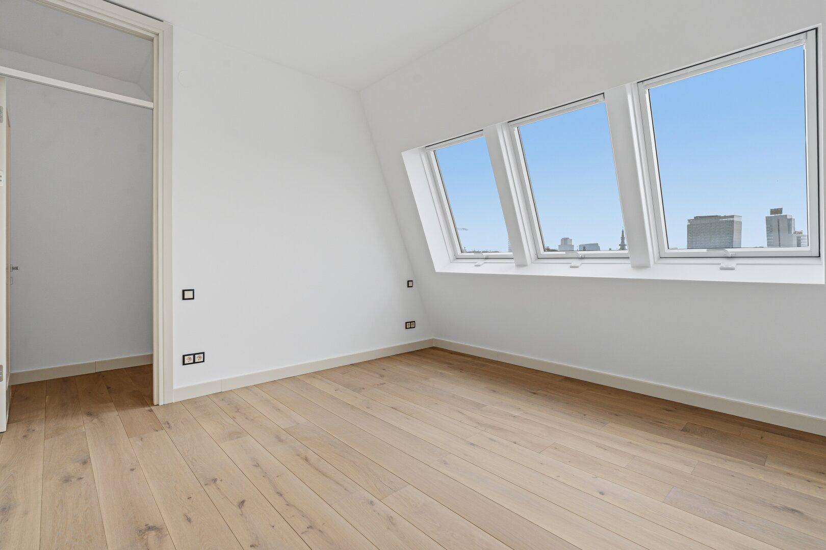 Lichtdurchflutetes Dachzimmer mit Holzboden und schrägen Fenstern, die einen Stadtblick ermöglichen.
