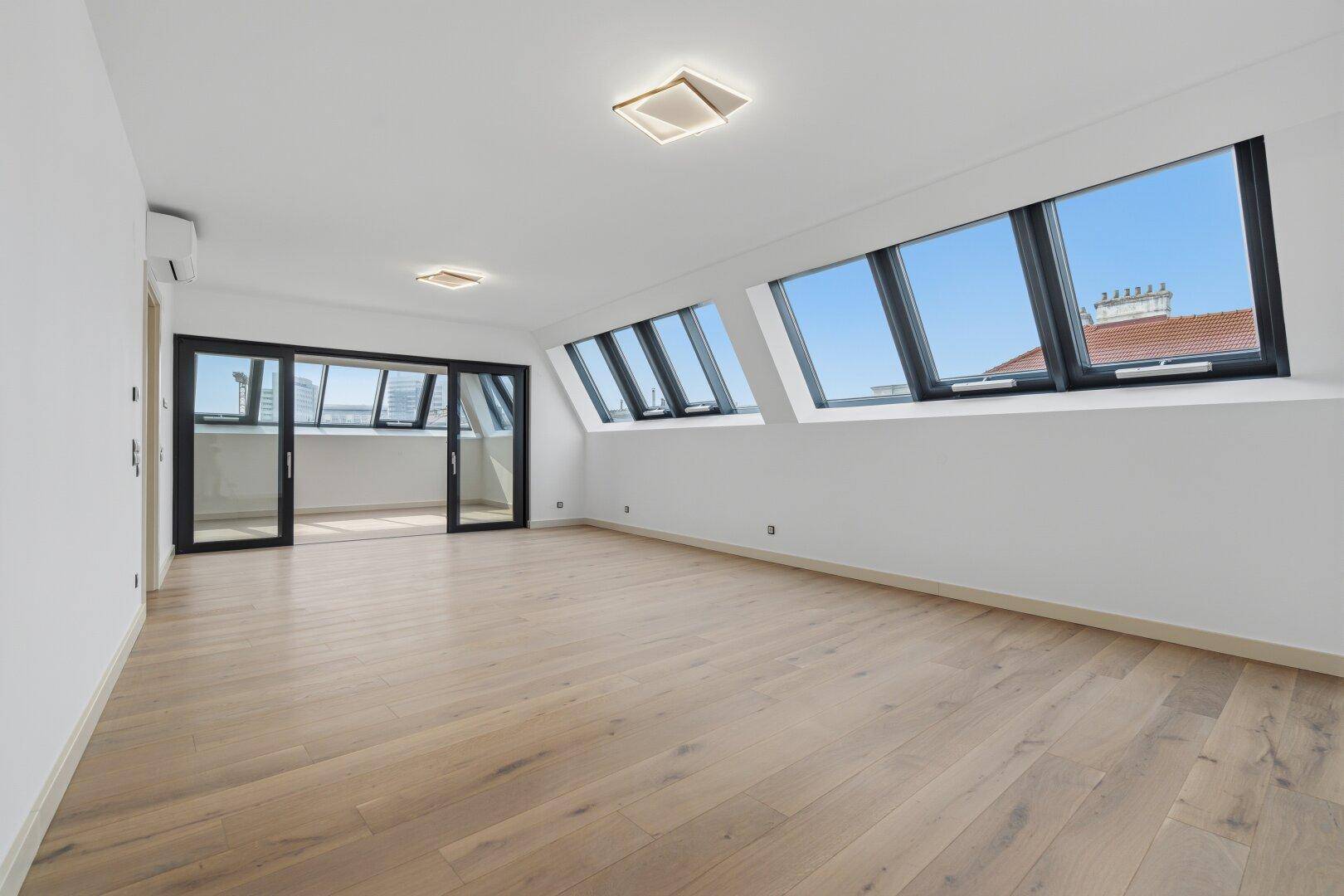 Heller, leerer Raum mit Dachschrägenfenstern und Glasschiebetüren, die zu einem Balkon führen.