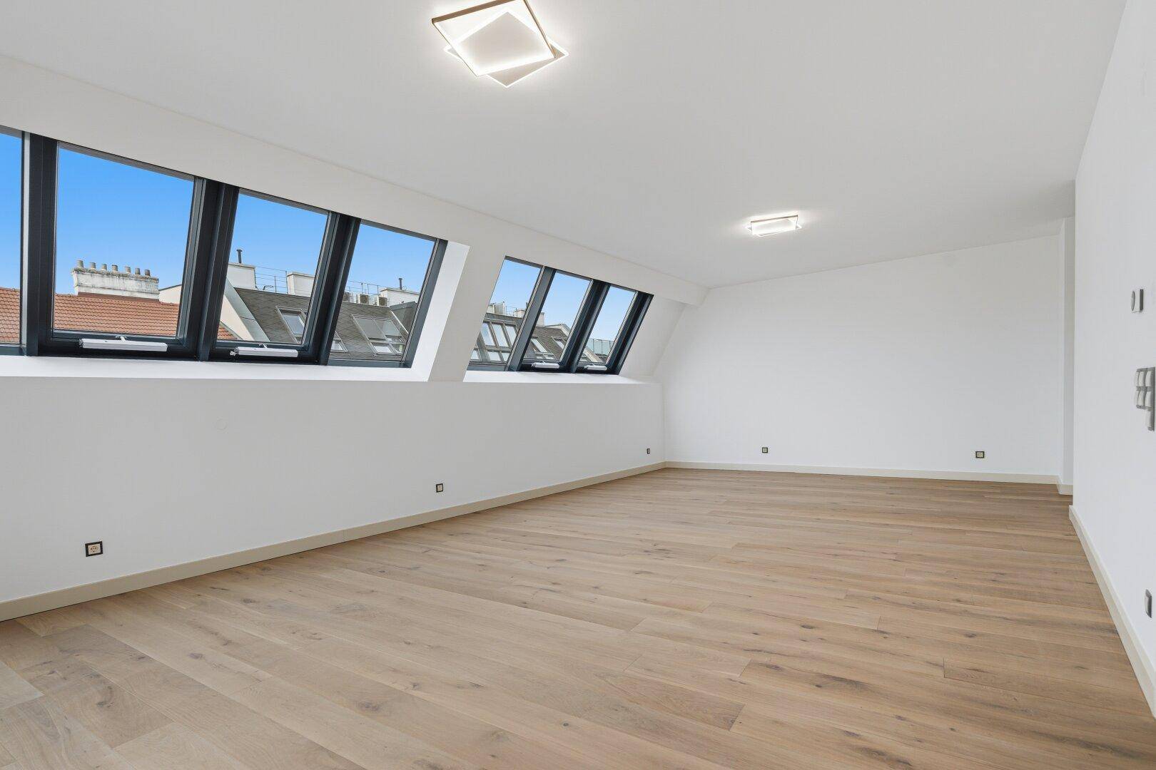 Heller, leerer Raum mit Holzboden und mehreren Dachschrägenfenstern für viel Tageslicht.