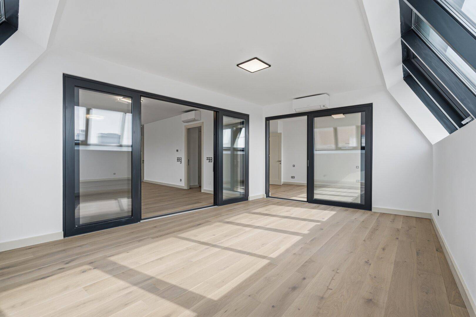 Großer, heller Raum mit Holzboden, Dachschrägenfenstern und mehreren Glasschiebetüren.