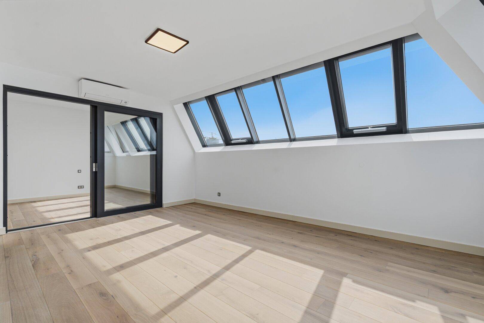 Heller, leerer Raum mit Holzboden, Dachschrägenfenstern und großer Glasschiebetür.