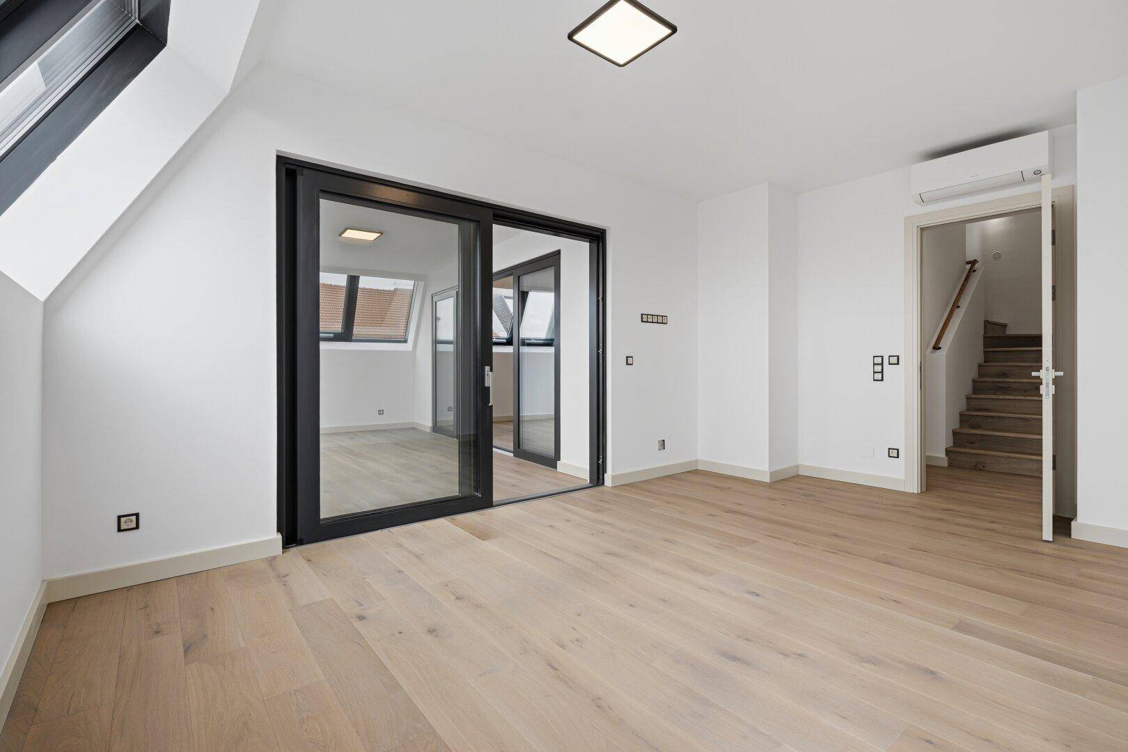 Heller Raum mit Holzboden, Glasschiebetüren und offenem Zugang zu einer Holztreppe.