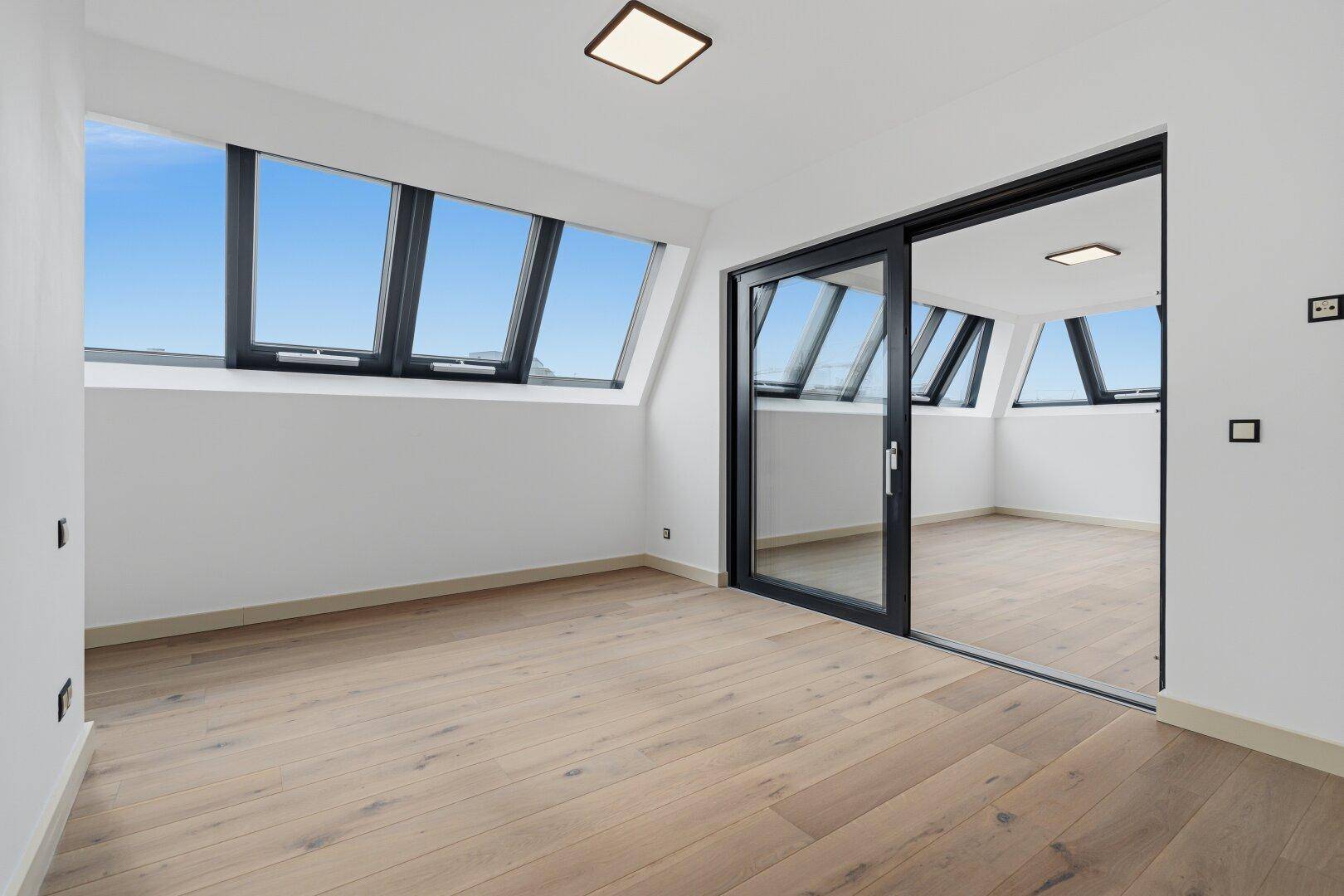 Heller, leerer Raum mit Holzboden, Dachschrägenfenstern und großer Glasschiebetür.