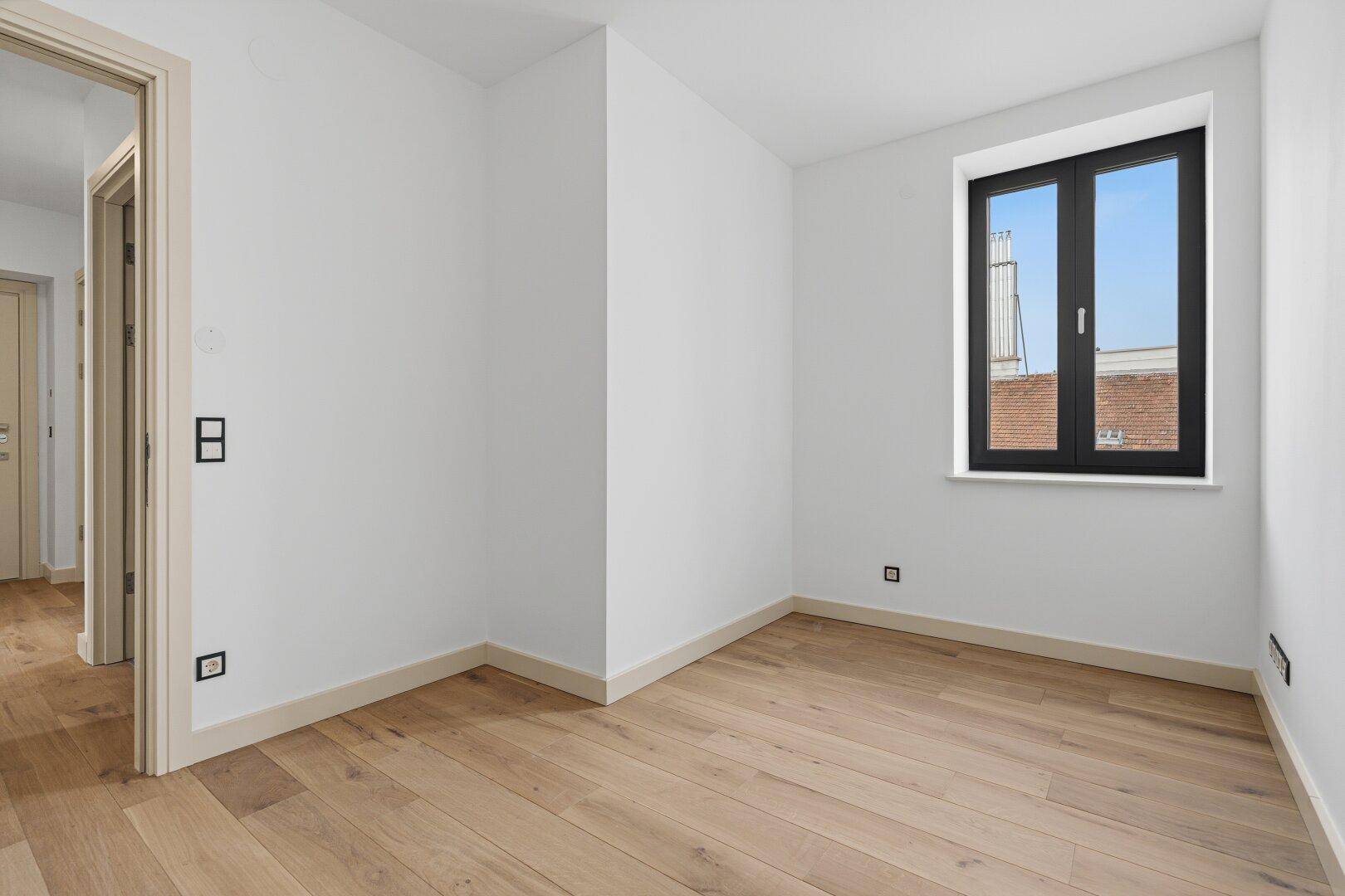 Helles, leeres Zimmer mit Holzboden und Fenster, bereit für individuelle Nutzung als Schlaf- oder Arbeitszimmer.