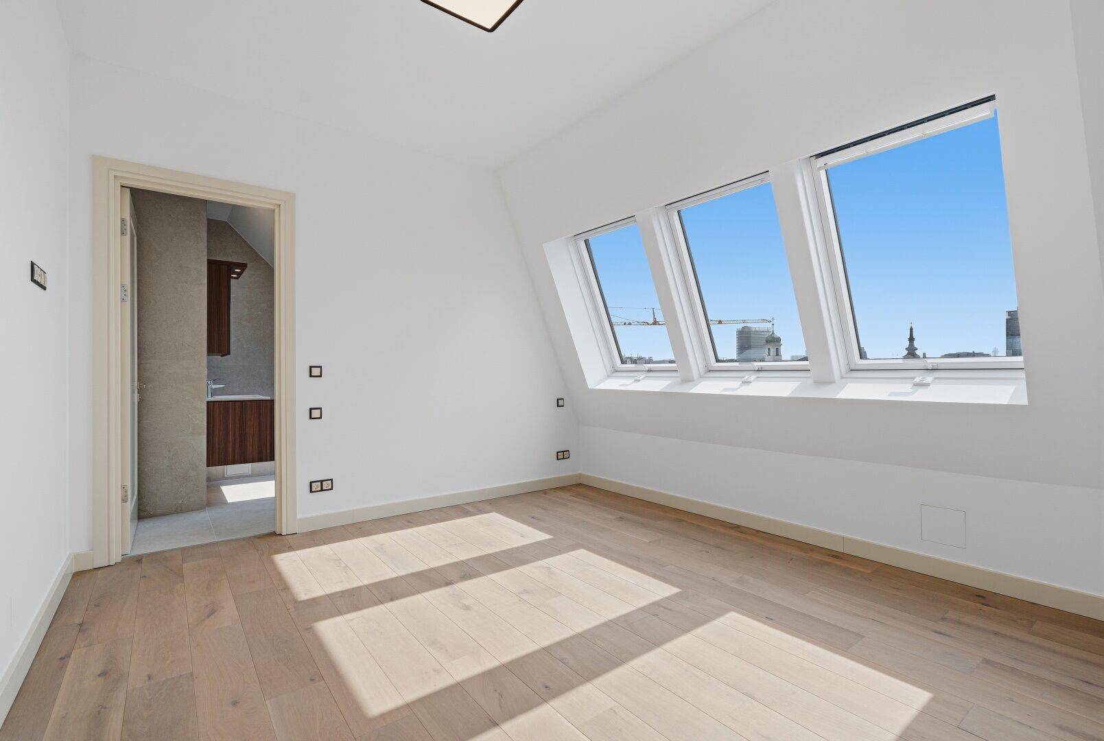Leeres Schlafzimmer mit Holzboden und Dachschrägenfenstern, Zugang zu einem angrenzenden Badezimmer.