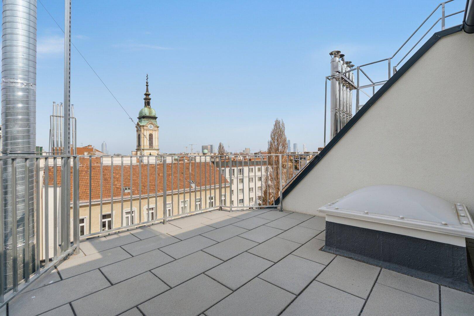 Geräumige Dachterrasse mit Stadtblick und einem markanten Kirchturm unter blauem Himmel.