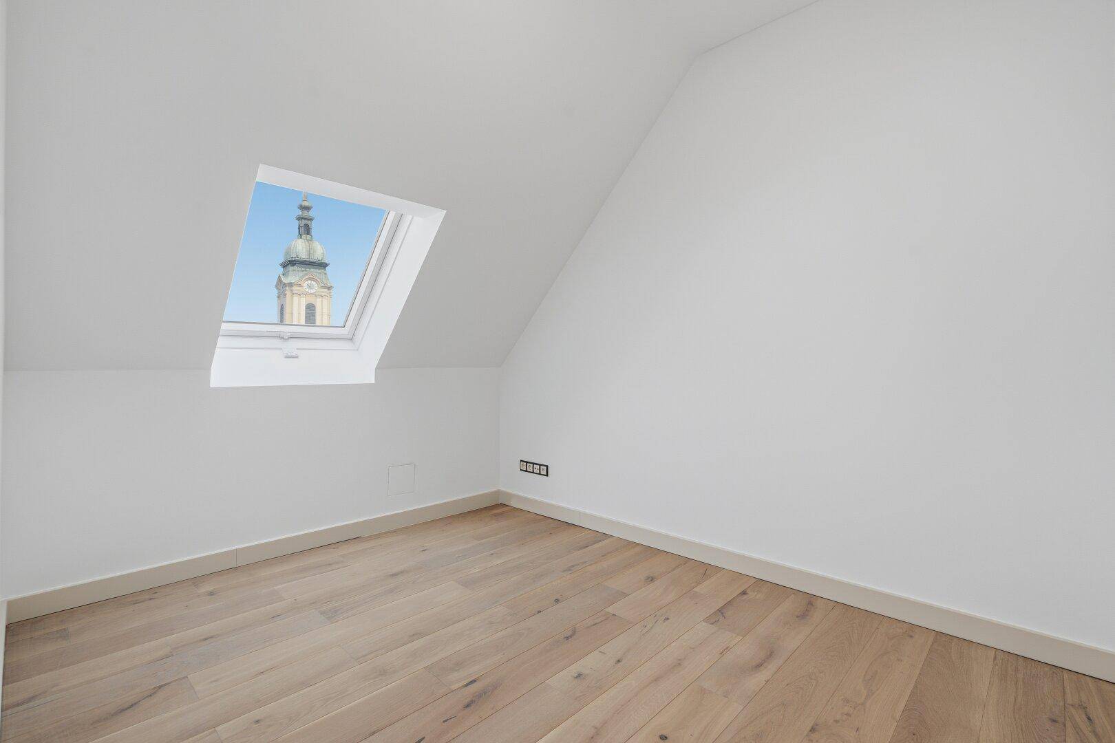 Leeres Zimmer mit Holzboden und Dachfenster, bietet einen Blick auf eine Kirchturmspitze.