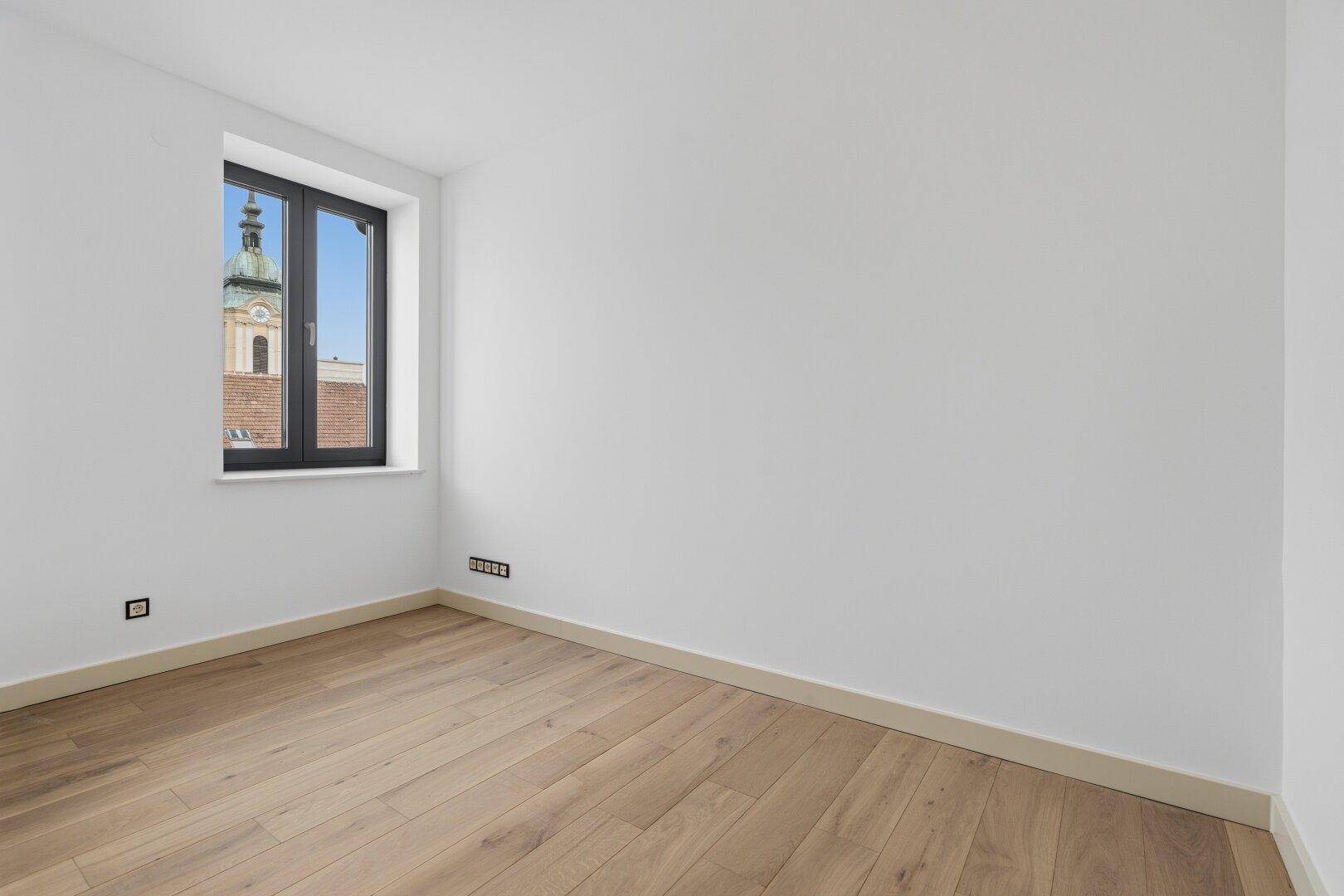 Leeres Zimmer mit Holzboden und Fenster, das einen Blick auf eine Kirchturmspitze bietet.