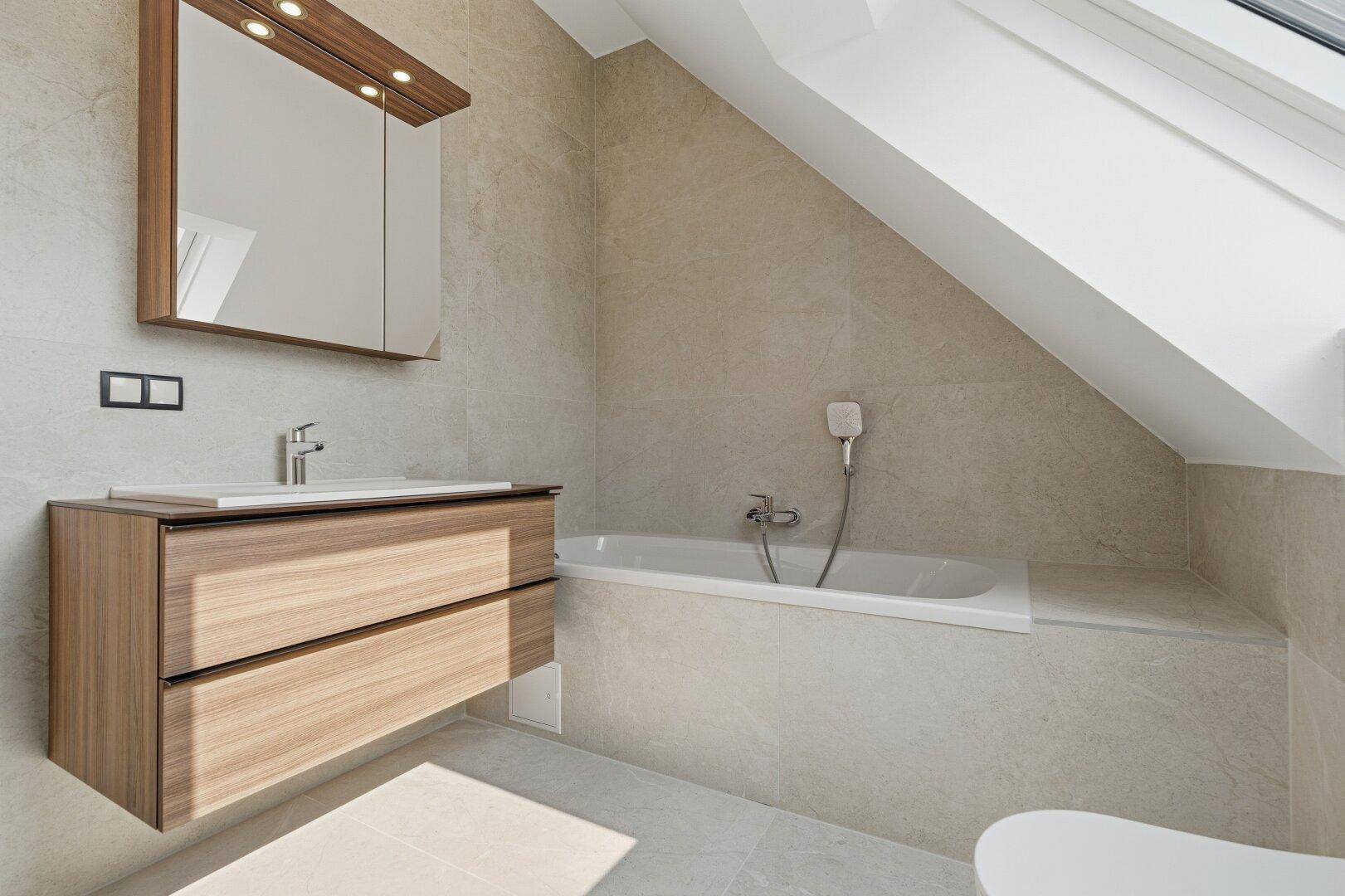 Helles Badezimmer mit Badewanne, Holzwaschtisch und Spiegelschrank, unter einem Dachfenster gelegen.