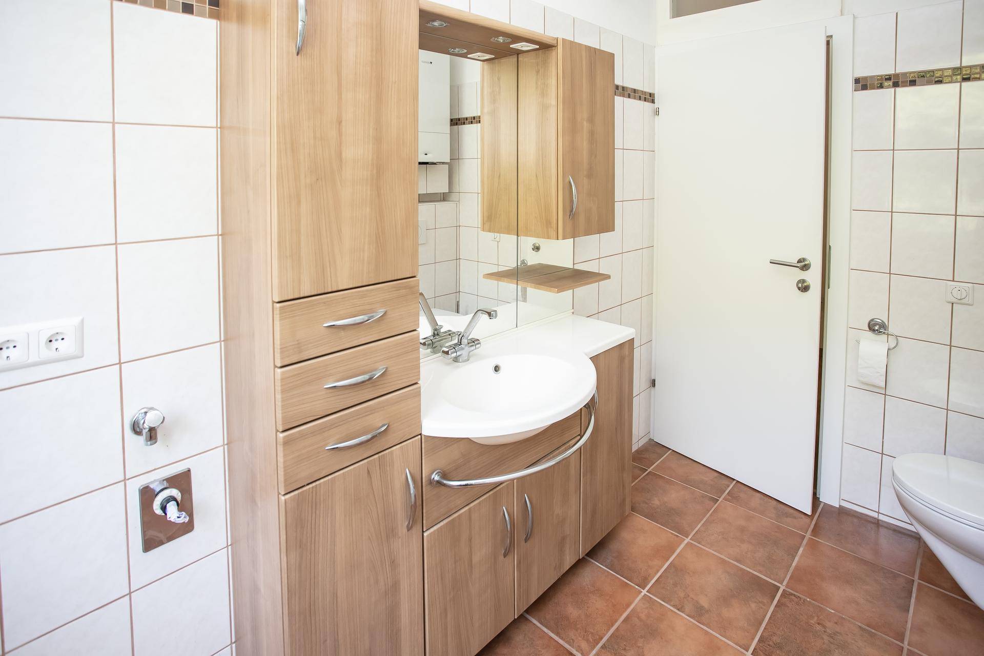 Badezimmer mit Holzschränken, weißem Waschbecken und hellen Fliesen.